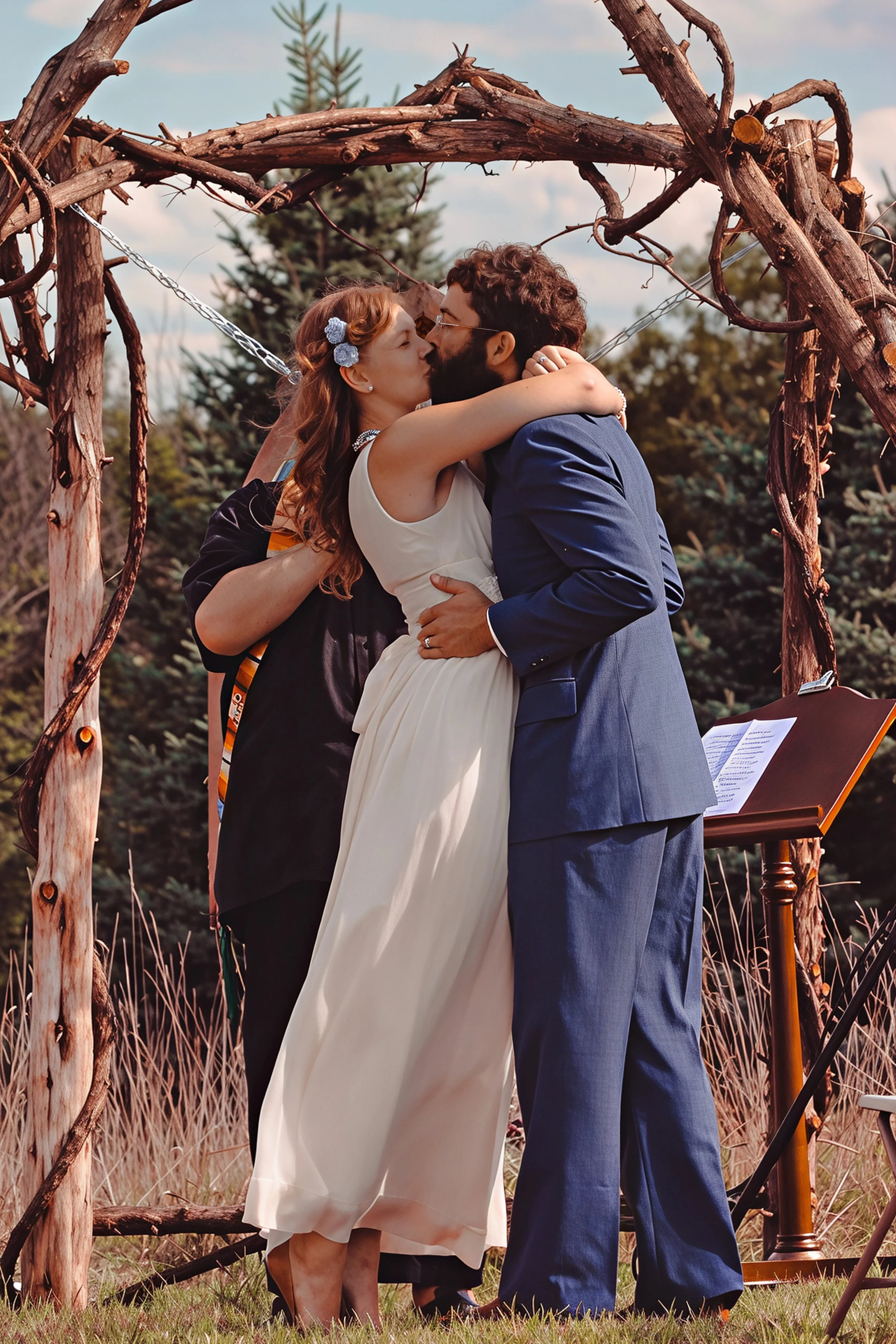 A bride and groom kissing during an outdoor wedding ceremony under a rustic wooden arch, with an officiant standing behind them, in front of a backdrop of trees and grass.