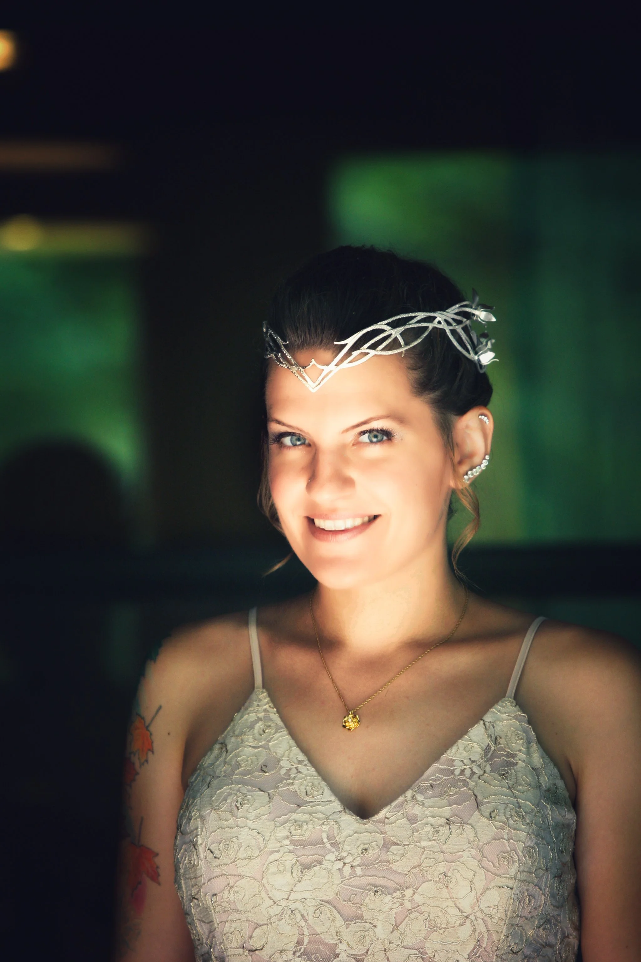 Portrait of a woman with a silver crown, blue eyes, and tattoos on her arm, smiling at the camera, wearing a lace top and gold necklace.