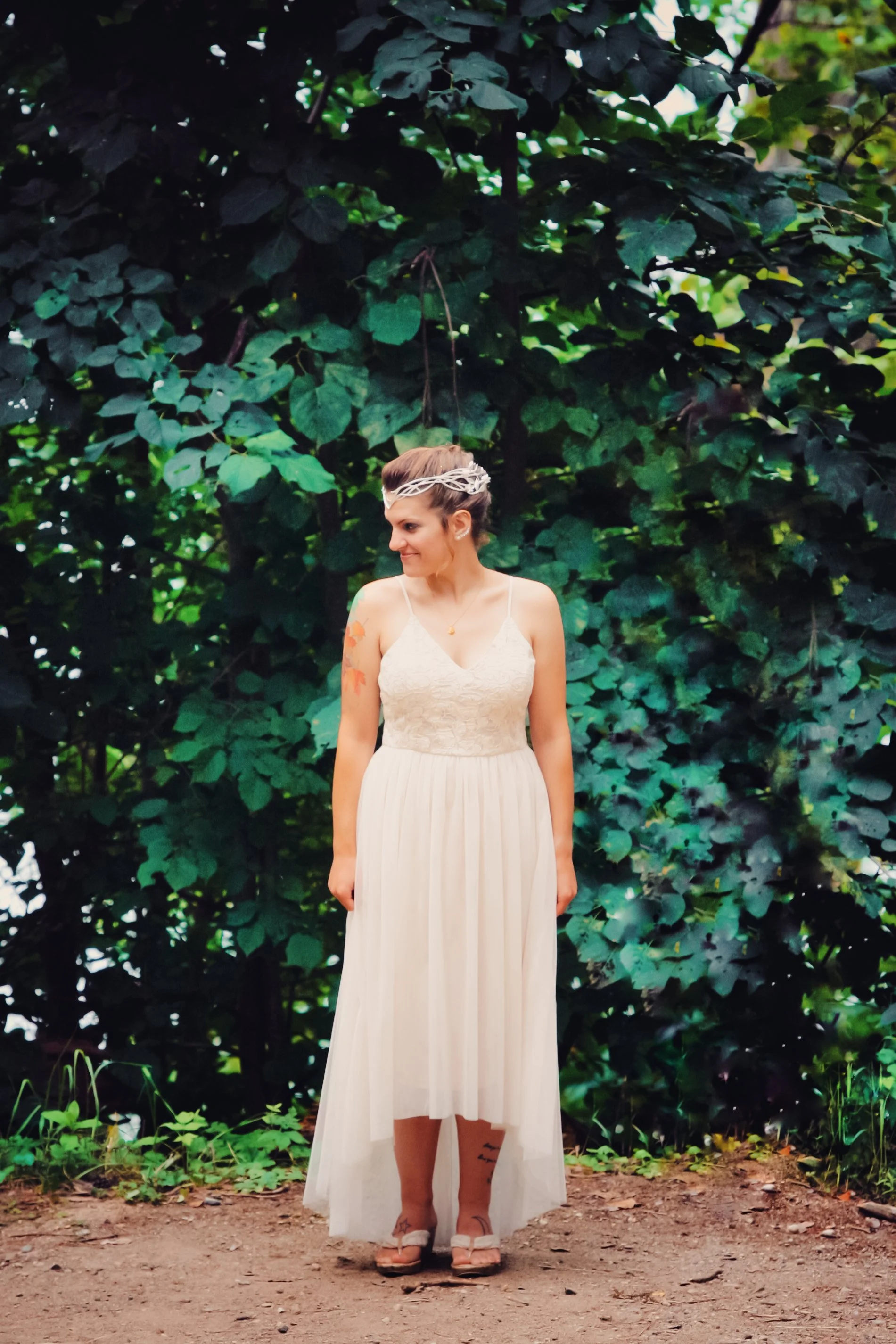 A woman in a white dress standing outdoors on dirt ground amidst lush green foliage, wearing a headband and sandals, looking to her right.