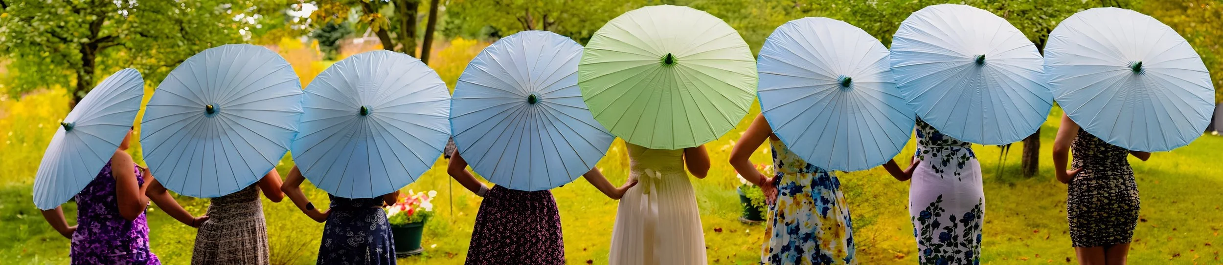 Group of women standing outdoors, holding blue and green umbrellas, wearing floral summer dresses in a park.