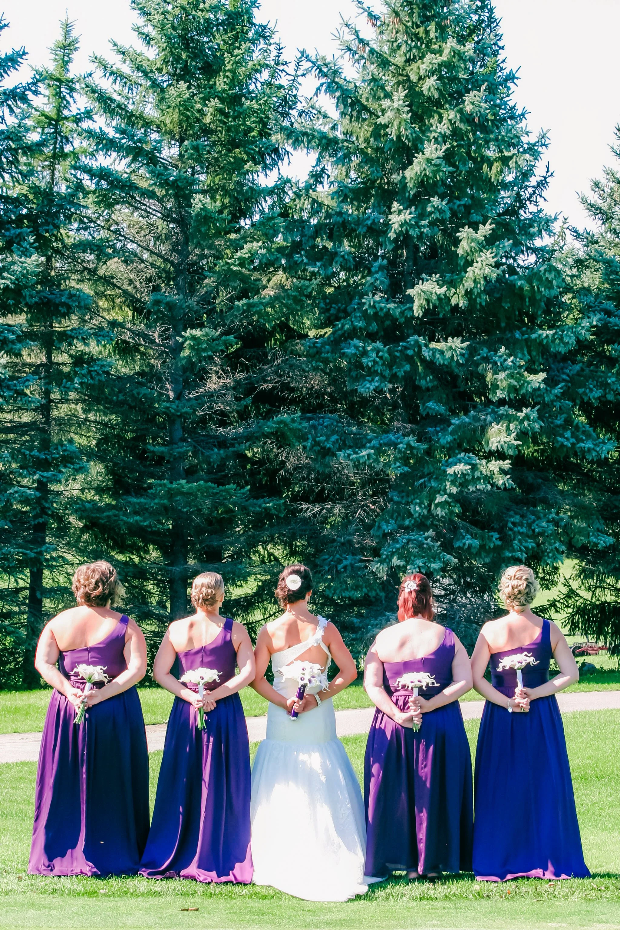 A bride standing with five women, likely bridesmaids, outdoors on a grassy area with large evergreen trees in the background. The bride wears a white wedding gown, and the bridesmaids wear matching purple dresses, all holding bouquets.