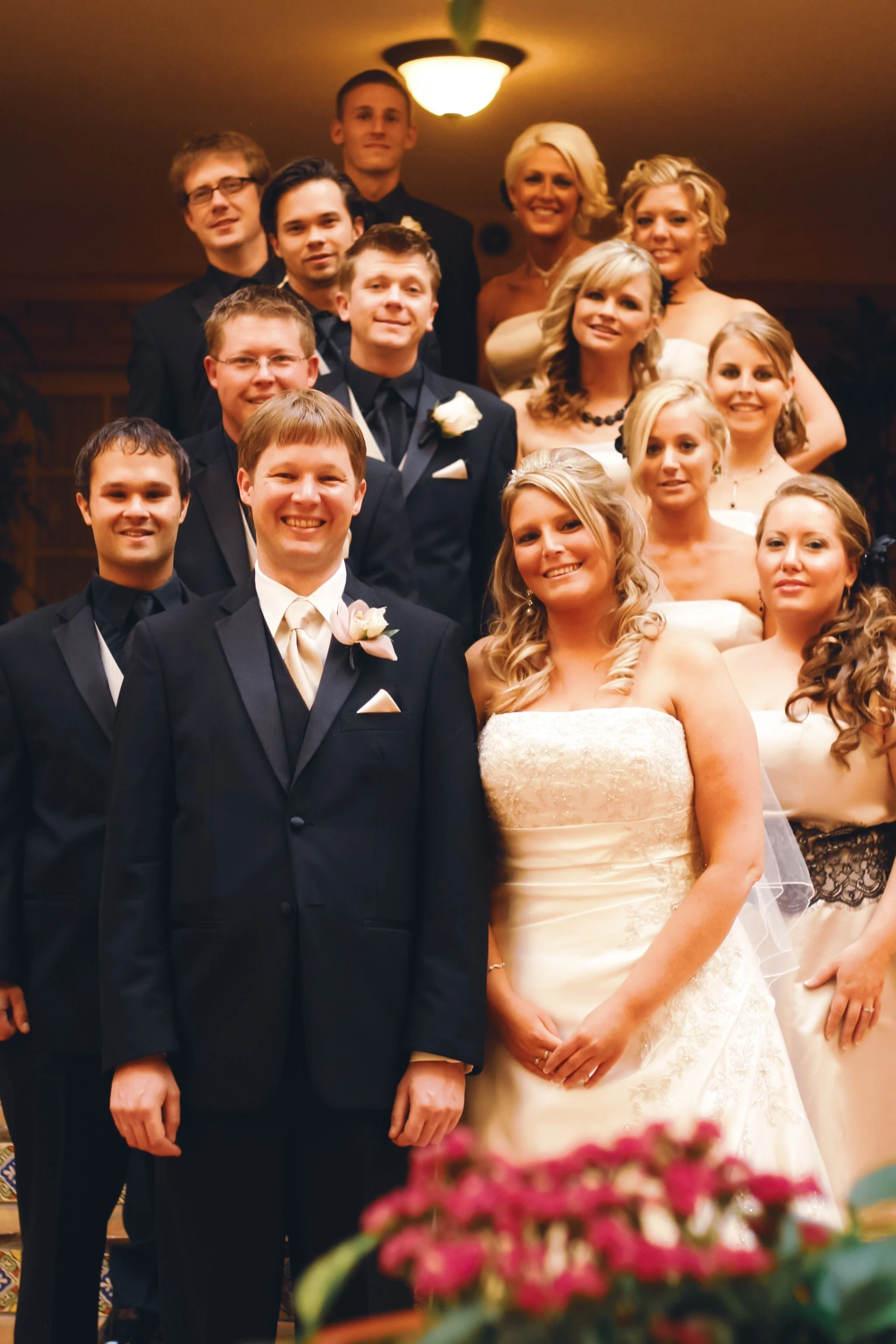 A wedding group photo including the bride and groom, accompanied by groomsmen and bridesmaids, posing indoors with warm lighting.