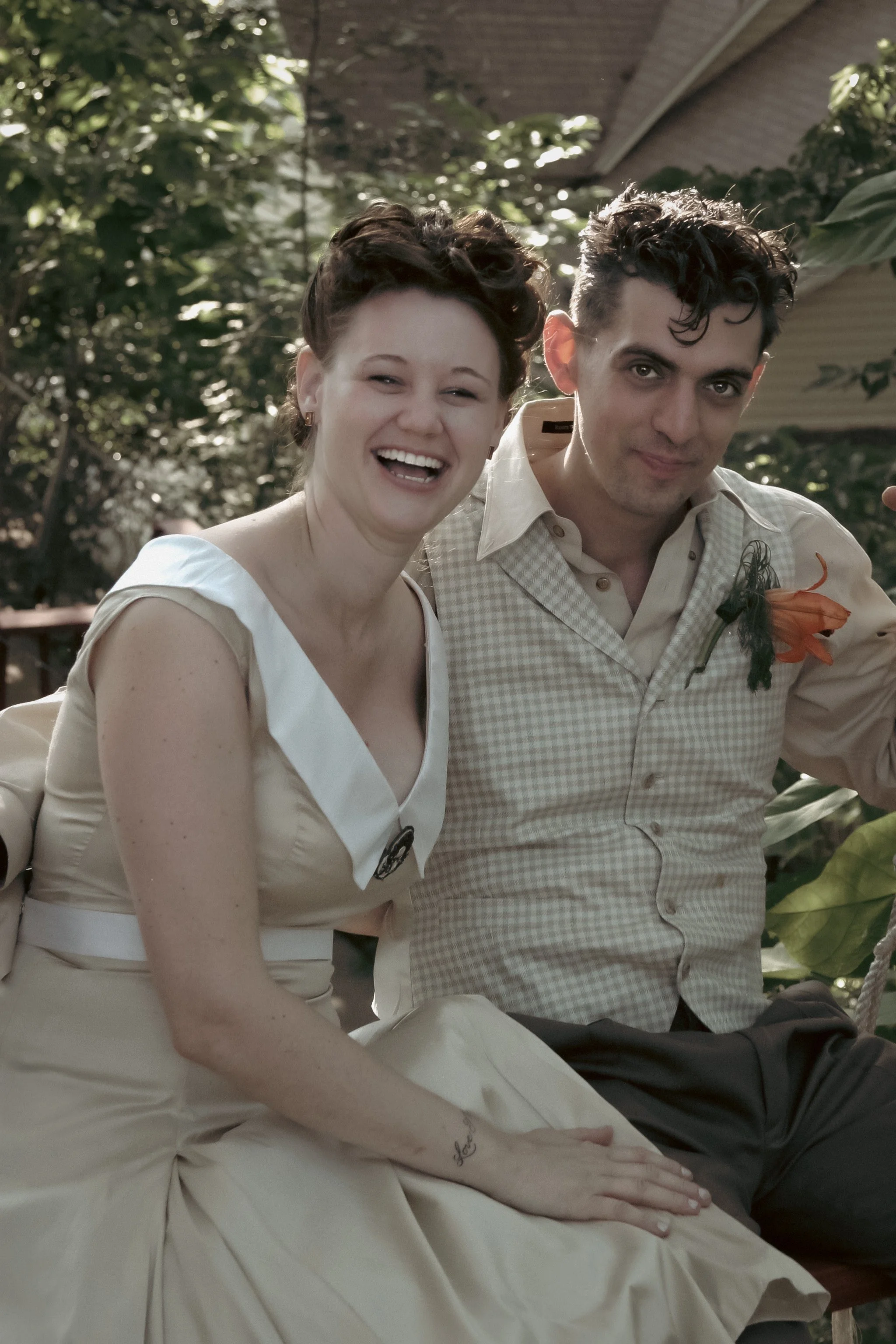 A woman and a man sitting together outdoors, smiling and looking at the camera. The woman is wearing a cream-colored dress with a white collar and the man is wearing a light-colored checkered shirt with a boutonniere.