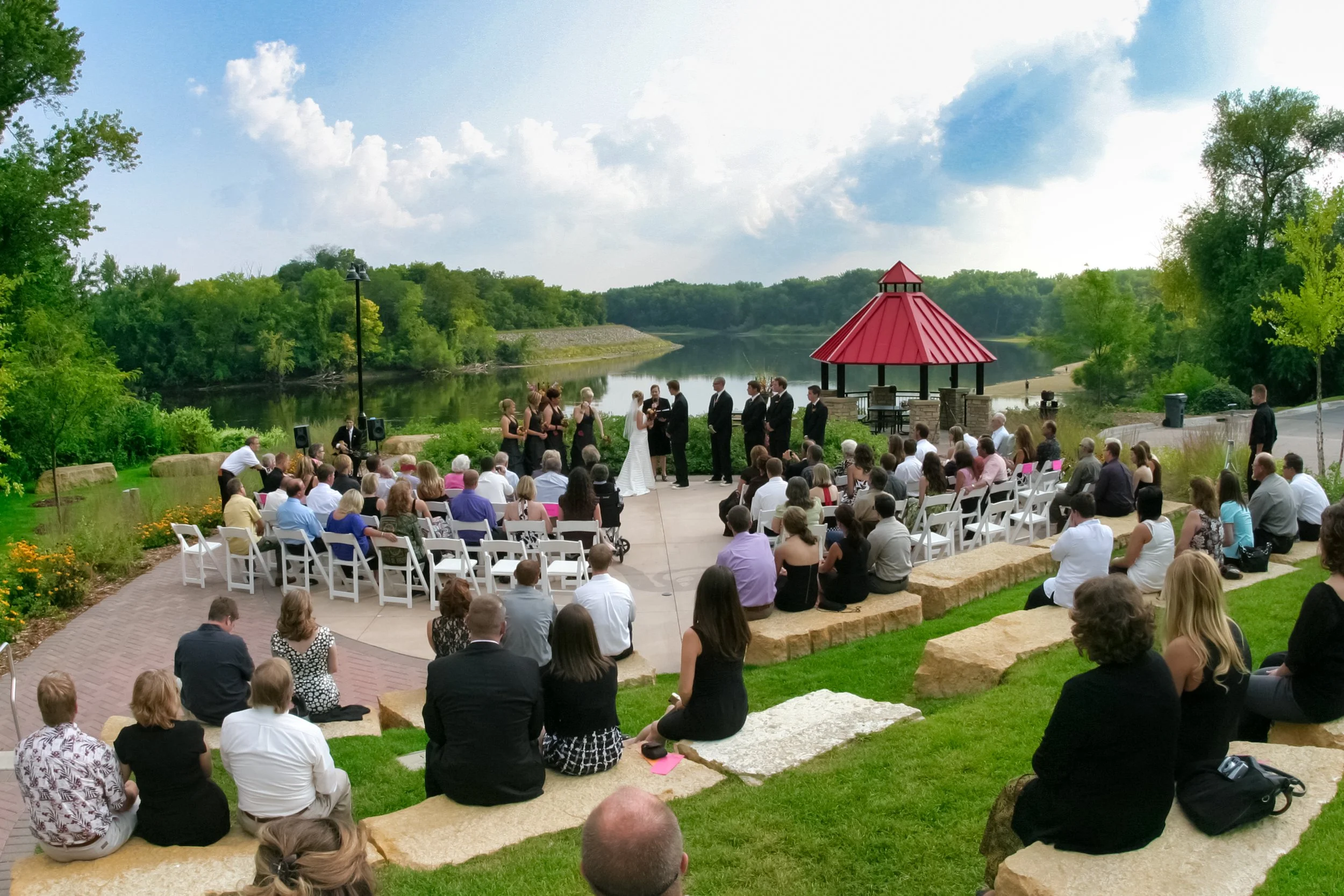 Outdoor wedding ceremony by a lake with guests seated on white chairs and on stone benches, a bride and groom exchanging vows, and a wedding party dressed in black and white, with green trees and a blue sky with clouds in the background.