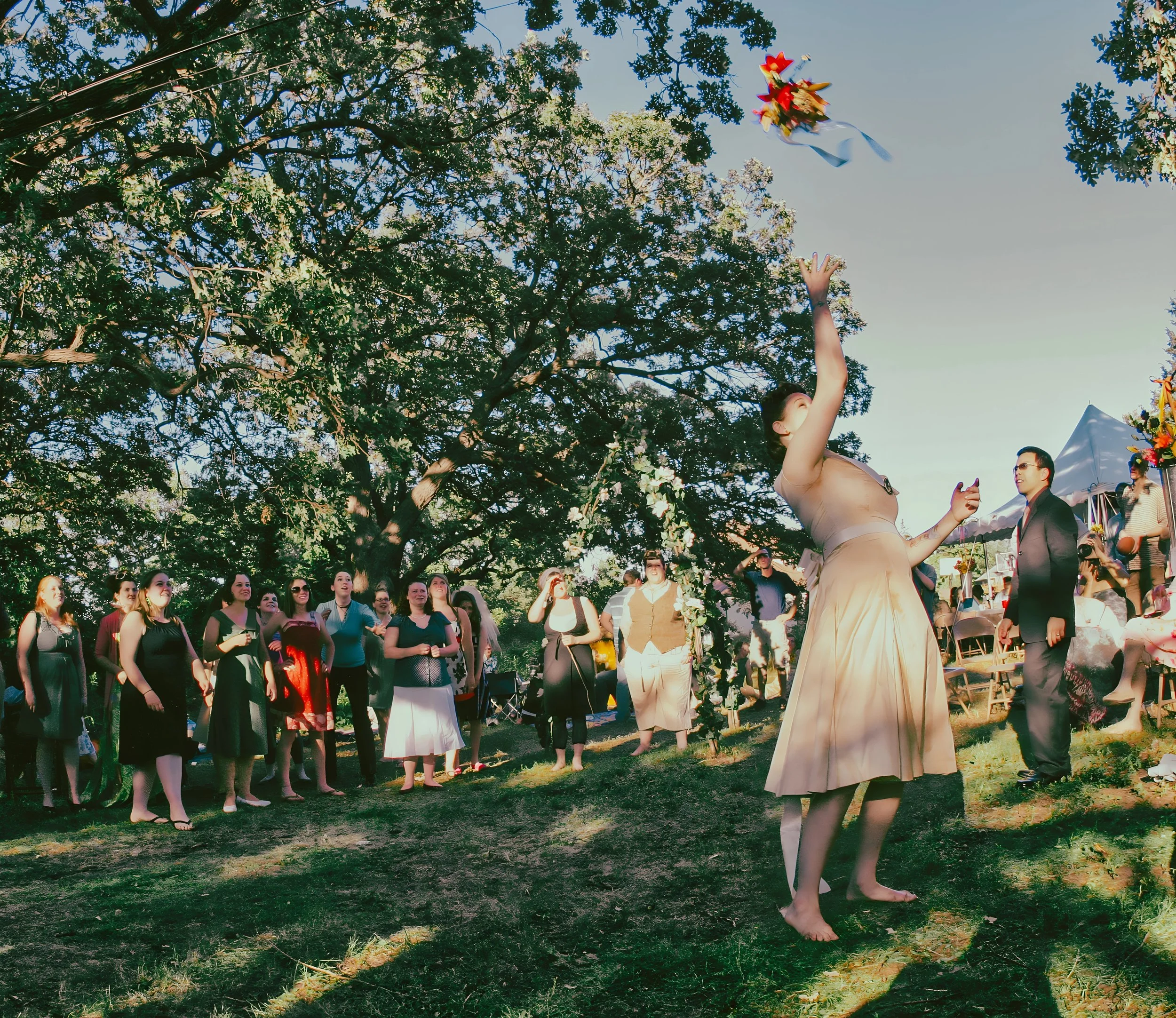 A woman in a beige dress is throwing a bouquet of flowers into the air at a wedding or outdoor event while a group of women stands in a line watching, under a large tree with green leaves. There are tents and other people in the background.