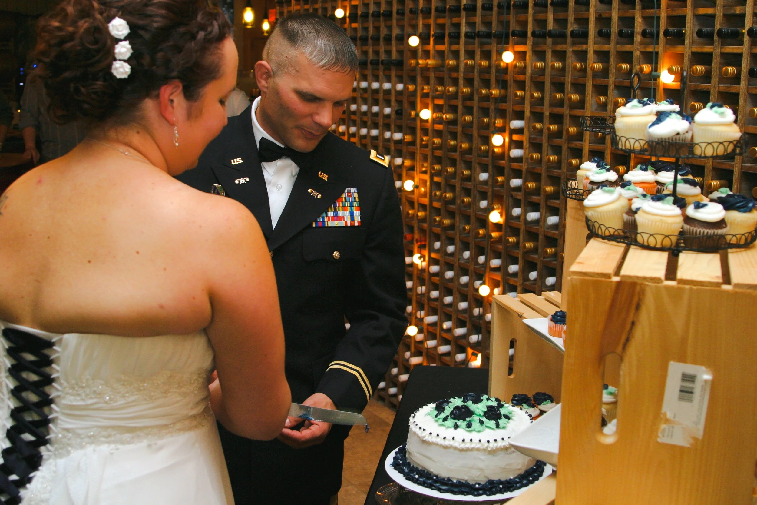 A bride and a groom in military uniform stand together at a wedding reception, looking at a cake. The background features a wine rack with bottles and warm lighting. There are cupcakes on display.