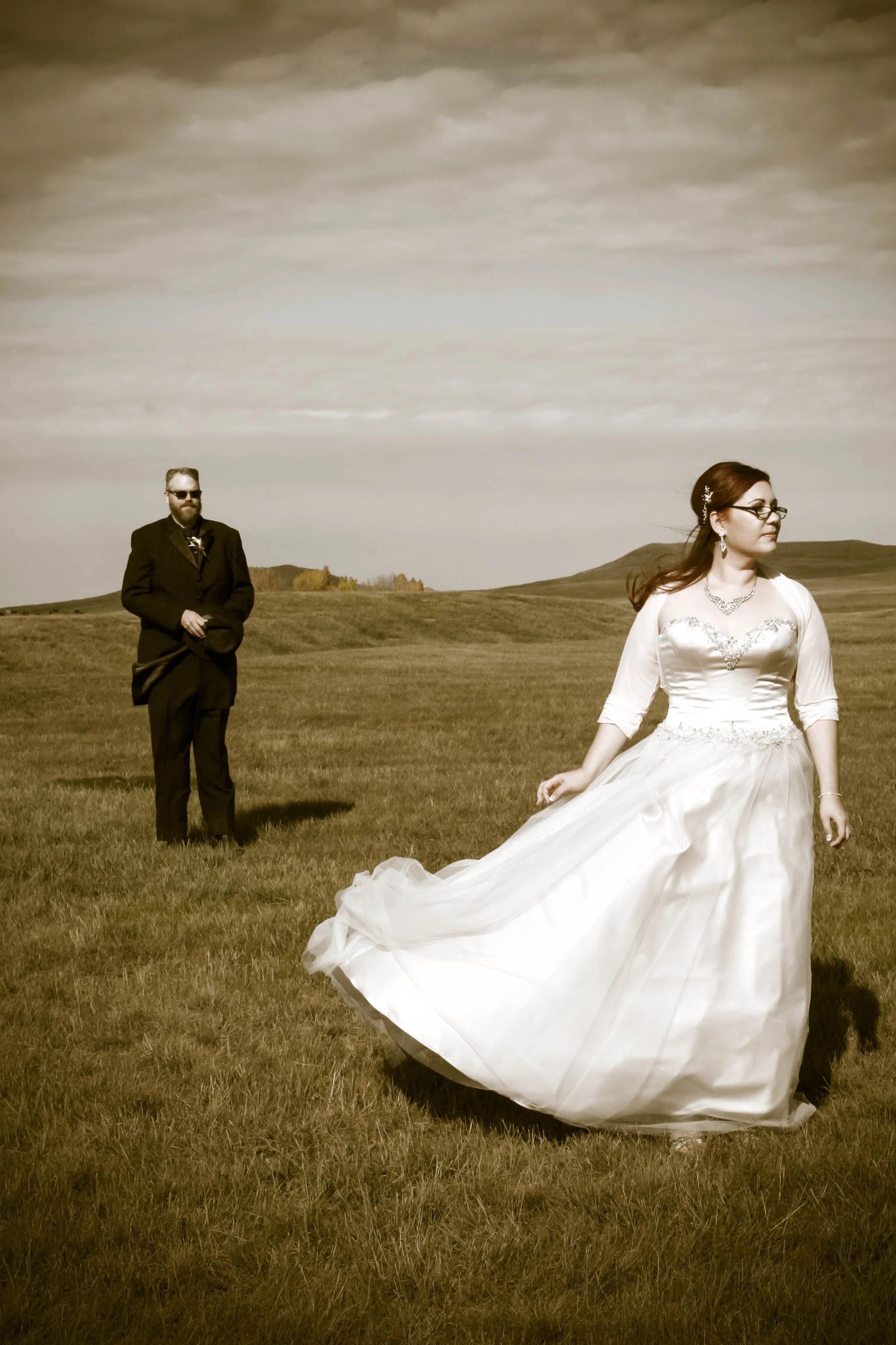 A bride in a white wedding dress standing in a grassy field with rolling hills in the background, a man in a suit standing further away, looking toward the bride, under a partly cloudy sky.