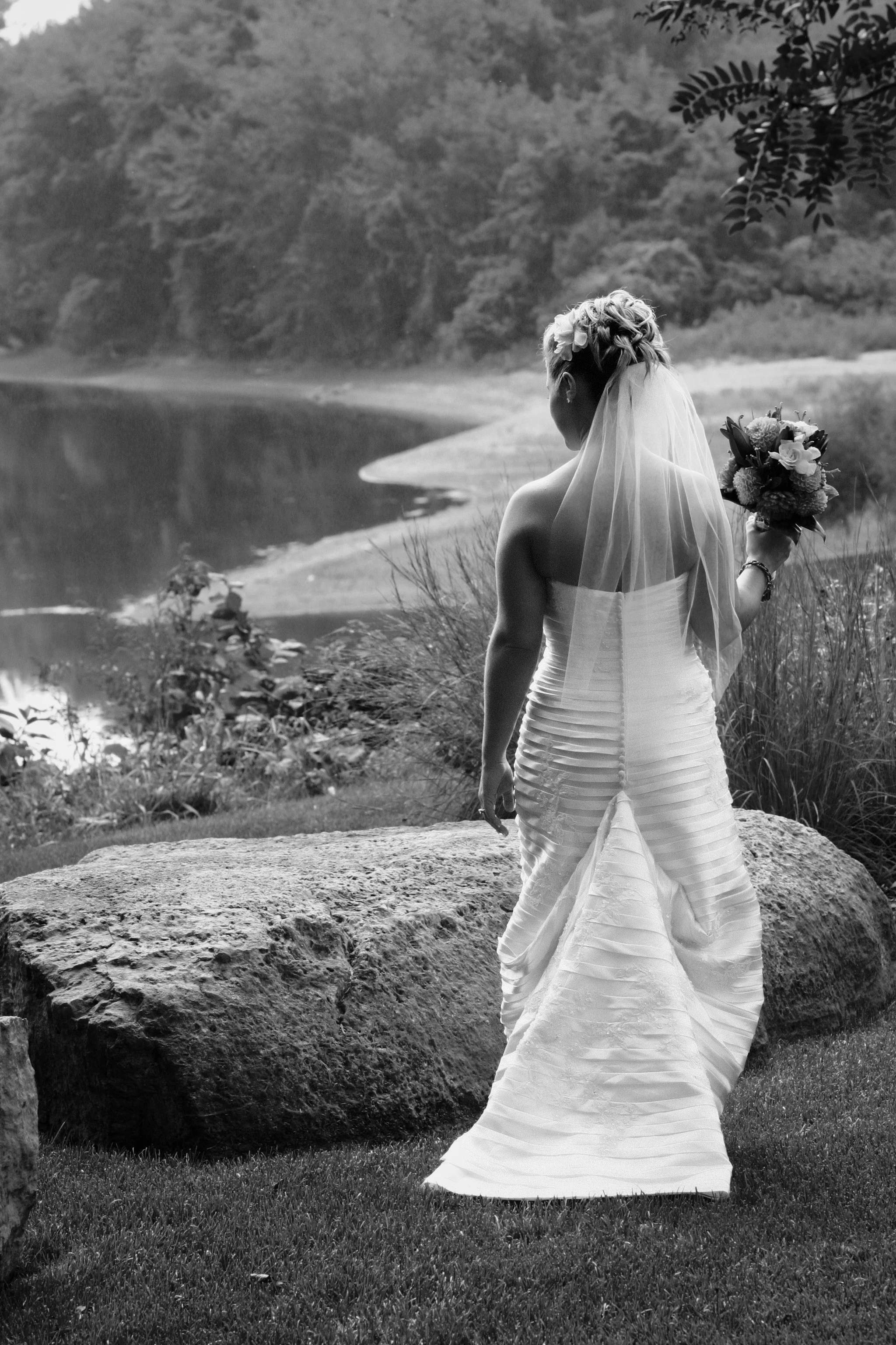 A bride in a strapless wedding gown with a long train, holding a bouquet of flowers, stands outdoors near a large rock, with a lake and trees in the background.