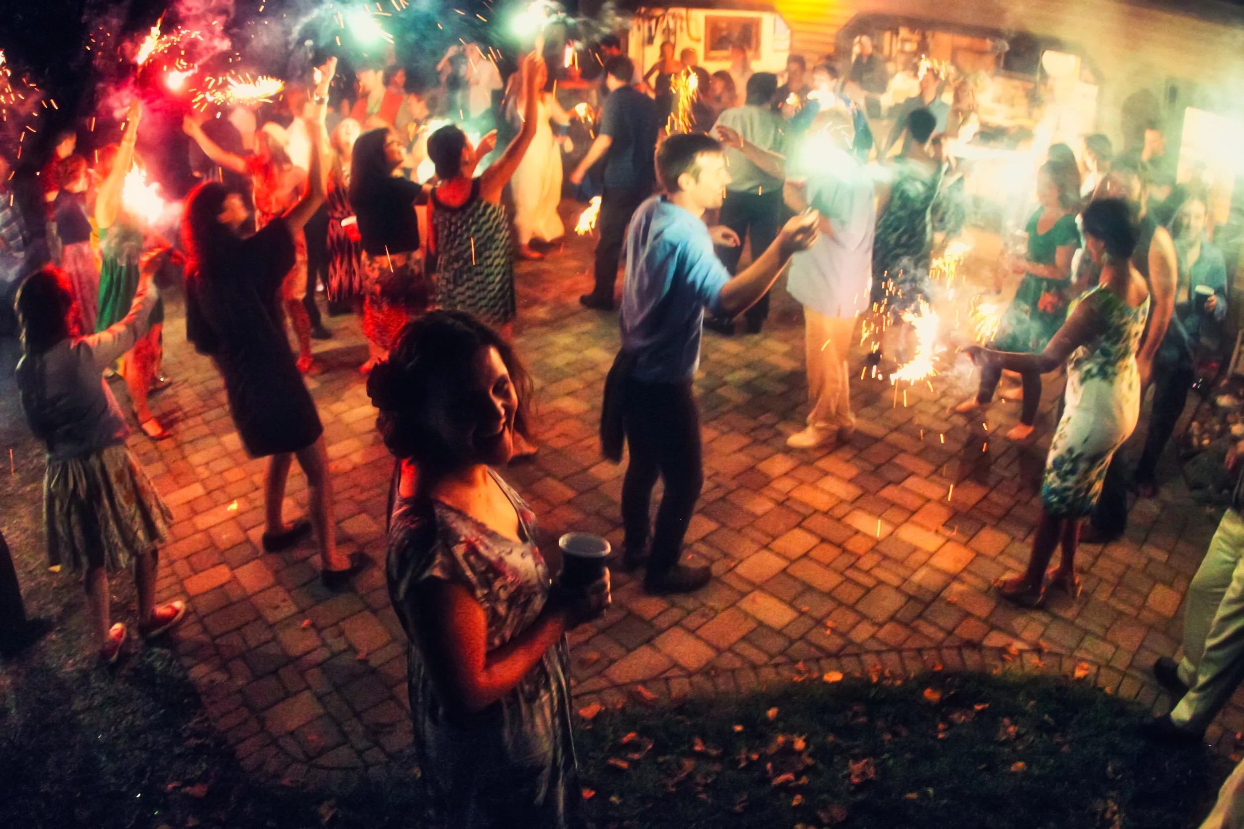 People celebrating outdoors at night with sparklers and fireworks, dancing and socializing.