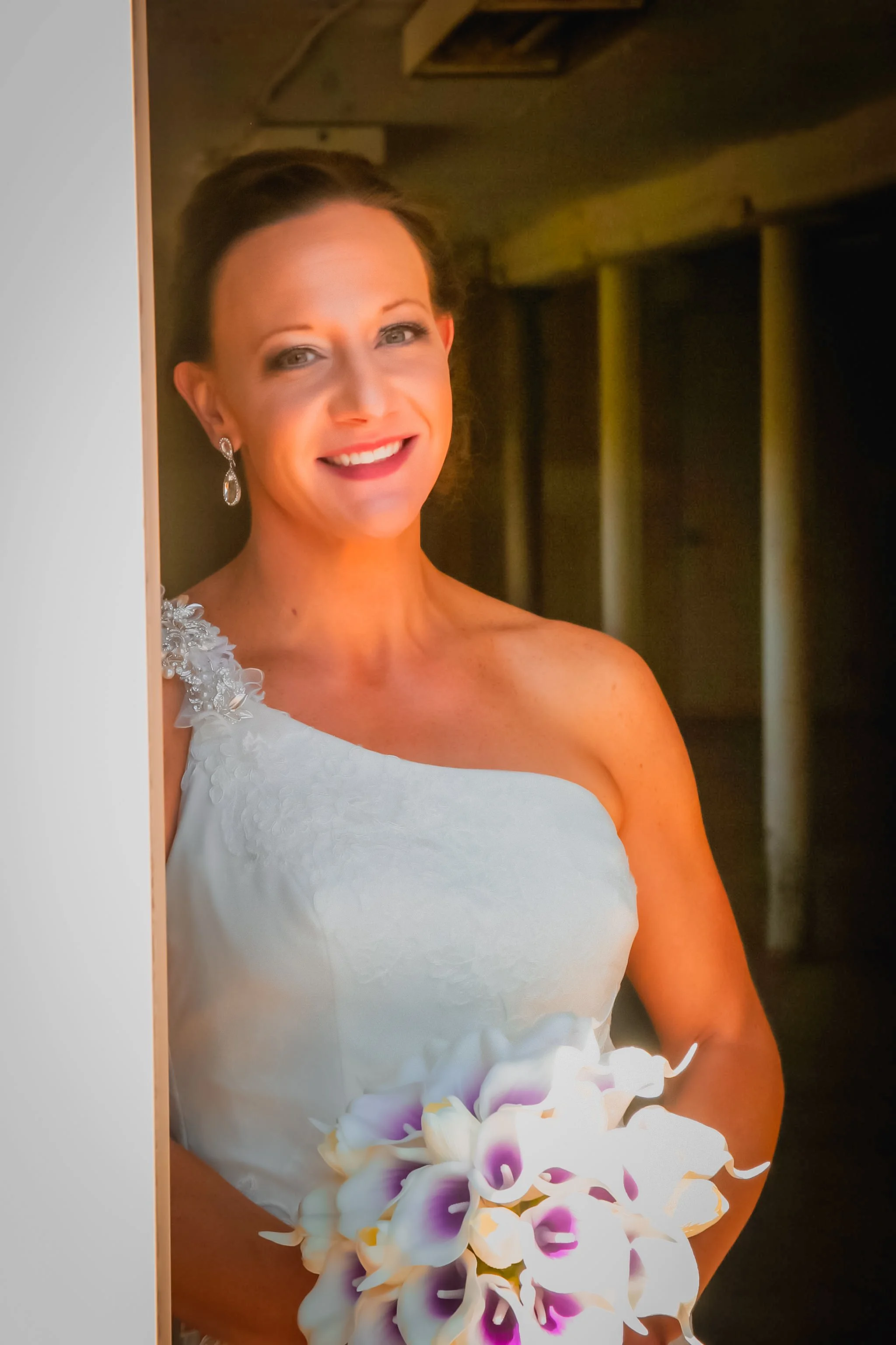 A smiling woman in a white dress holding a bouquet of white and purple orchids, standing in a dimly lit indoor setting.