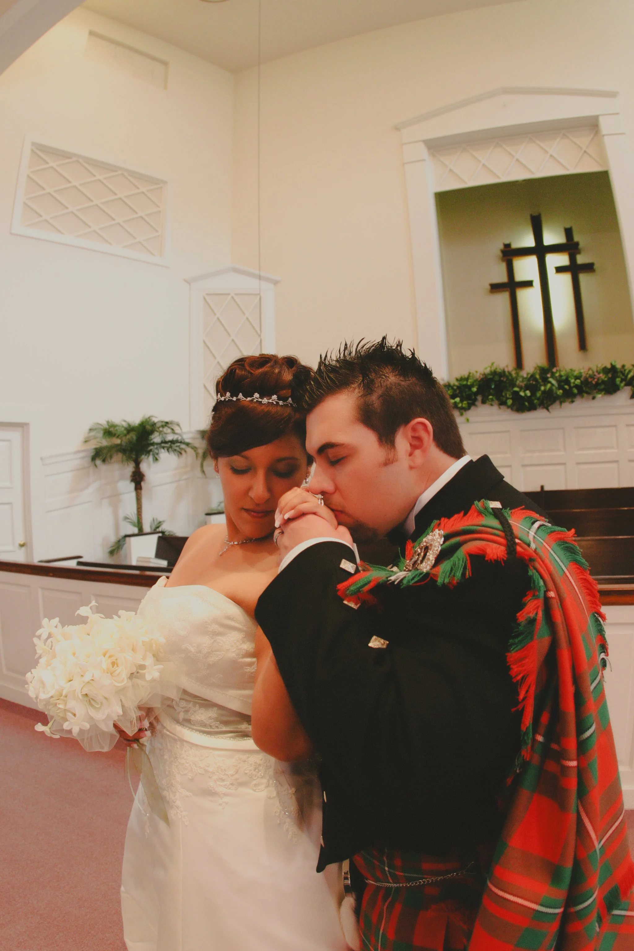 A bride and groom sharing a moment of prayer or reflection inside a church, with the bride holding a bouquet and the groom dressed in traditional Scottish attire, including a tartan kilt and sash.