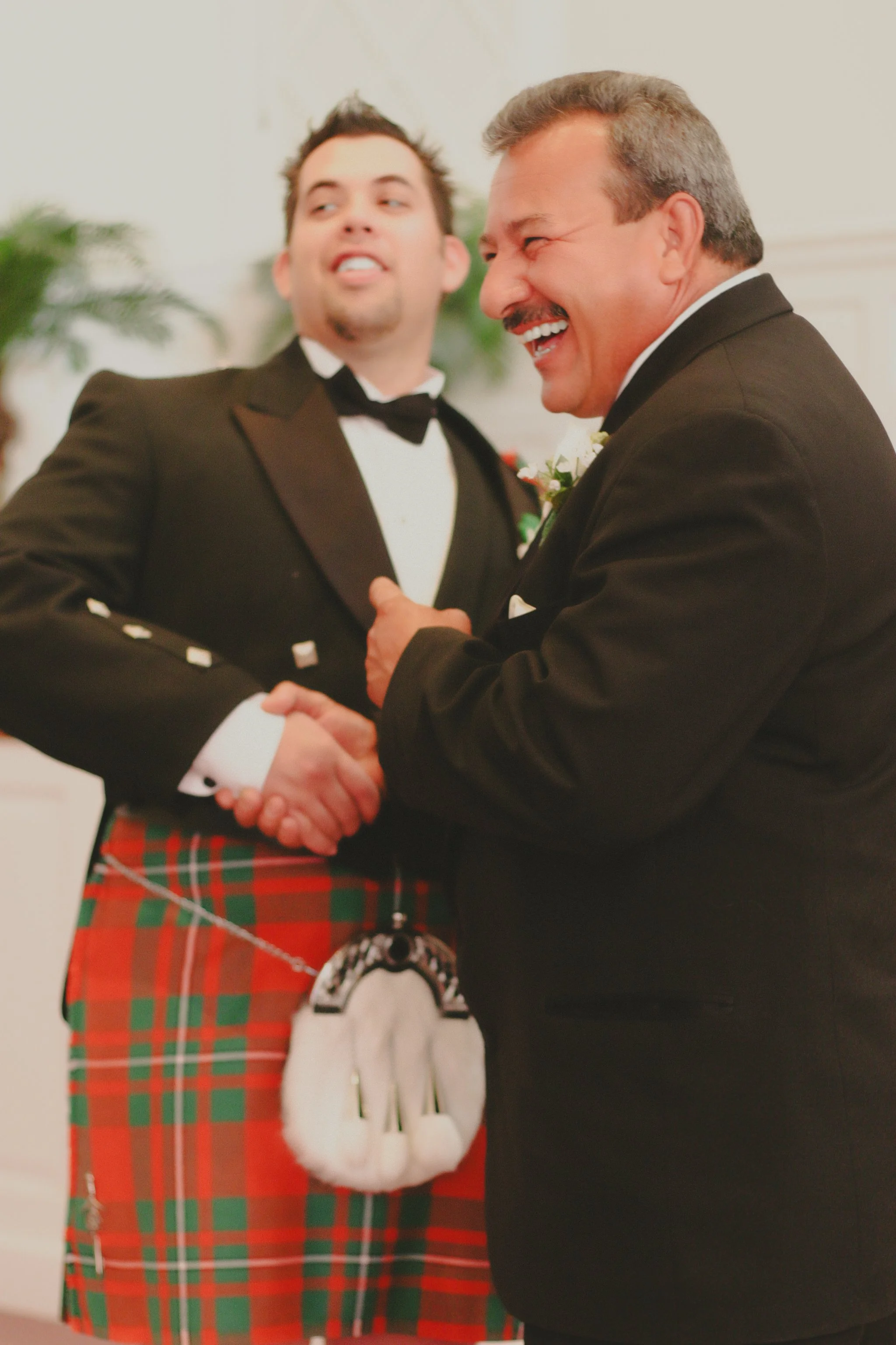 Two men in tuxedos shaking hands at a formal event, one wearing a tartan kilt and sporran, both smiling and laughing.