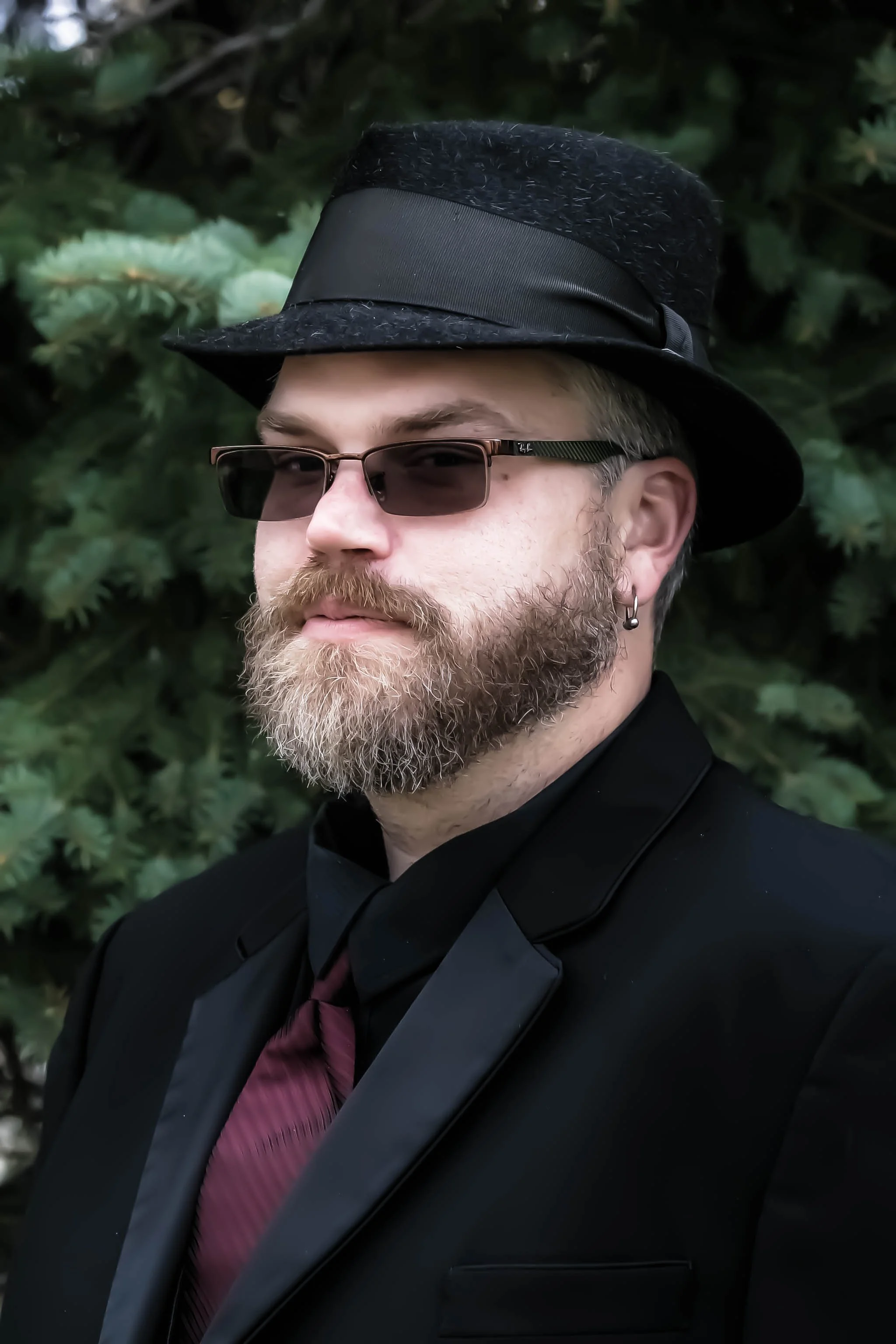 A man with a beard wearing sunglasses, a black suit, a purple tie, a black fedora hat with a black band, and an earring, standing in front of a leafy background.