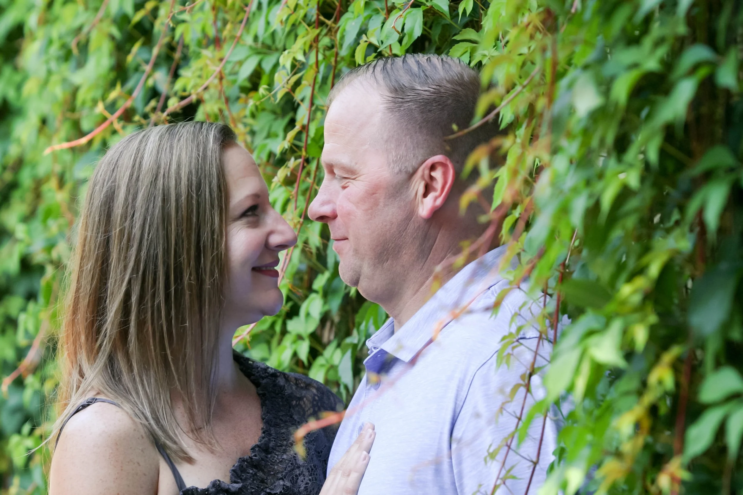 A couple standing close to each other, facing each other with smiles, surrounded by green leafy vines.