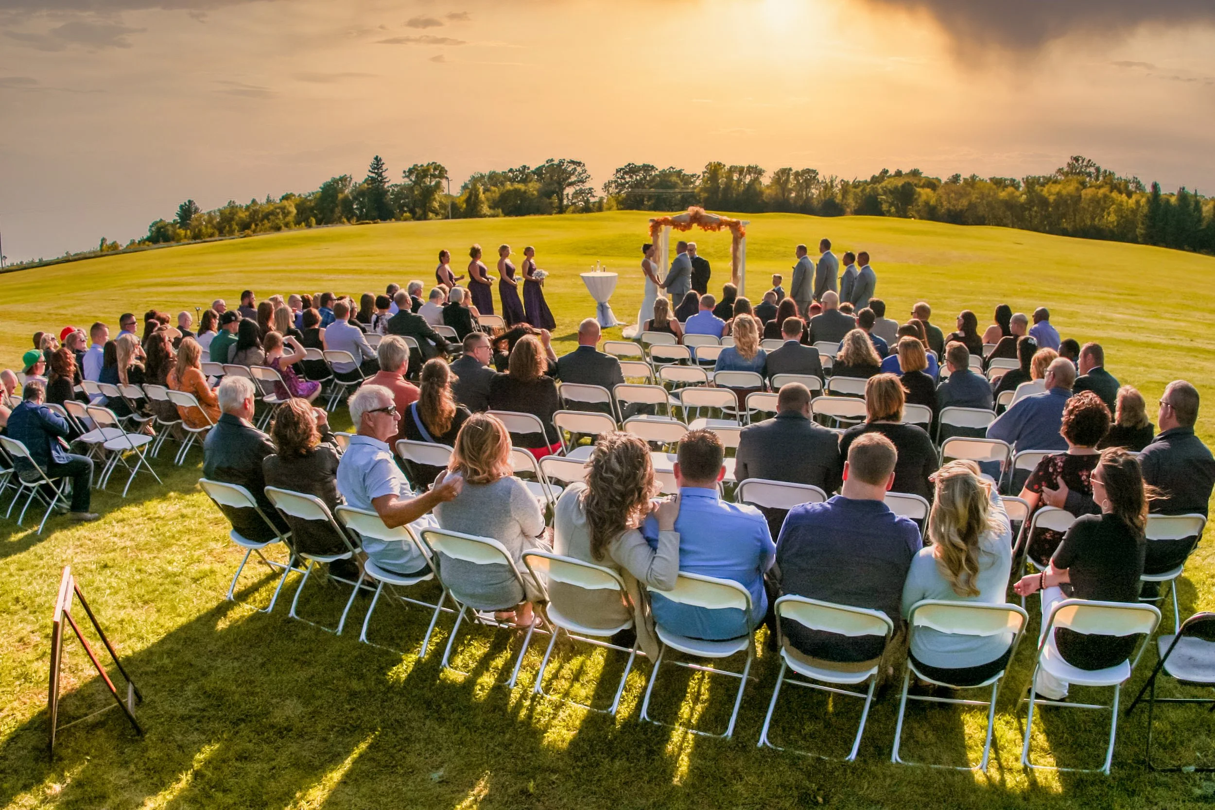 Outdoor wedding ceremony with guests seated on white chairs on a grassy lawn, officiants and couple under a floral arch, during sunset.