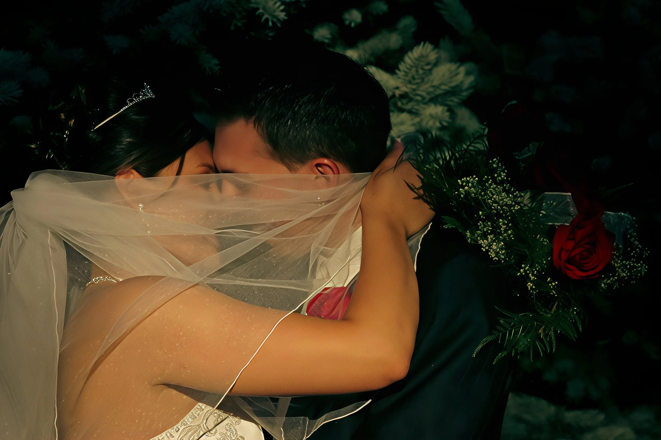 A bride and groom are embracing during their wedding, with the bride wearing a veil and a tiara, holding a bouquet of red roses and greenery.