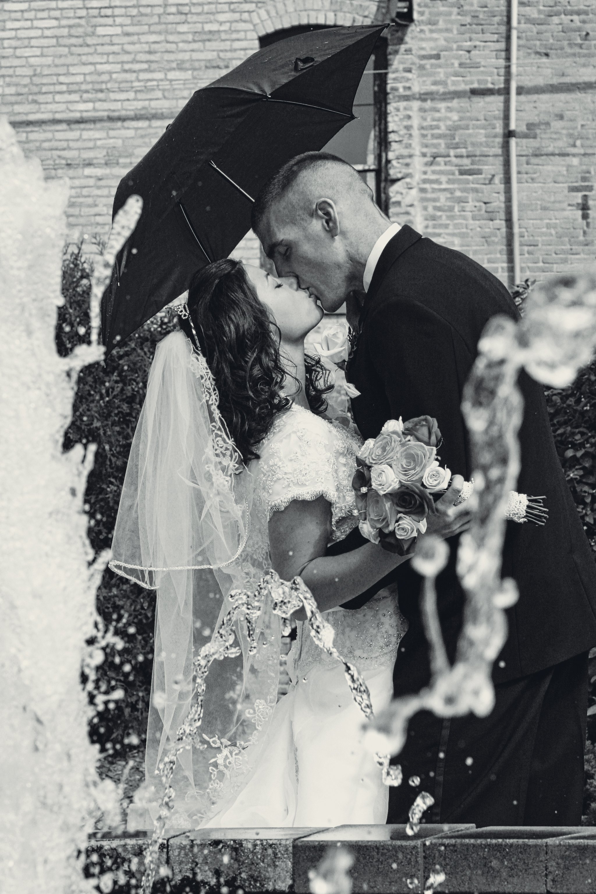A black and white photo of a newlywed couple sharing a kiss under an umbrella, with water splashing around them, in front of a brick building.