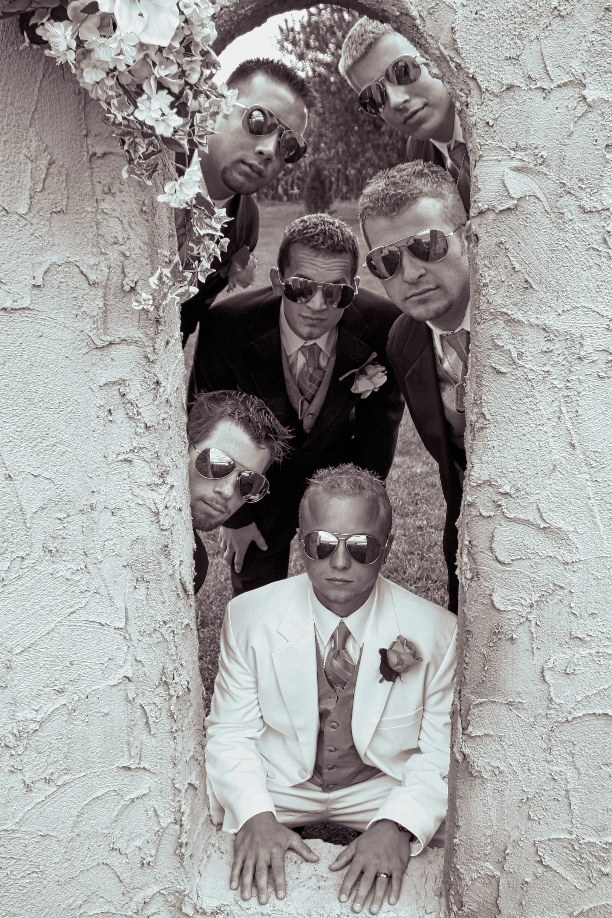 Men dressed in suits and sunglasses looking through a circular opening in a textured wall.
