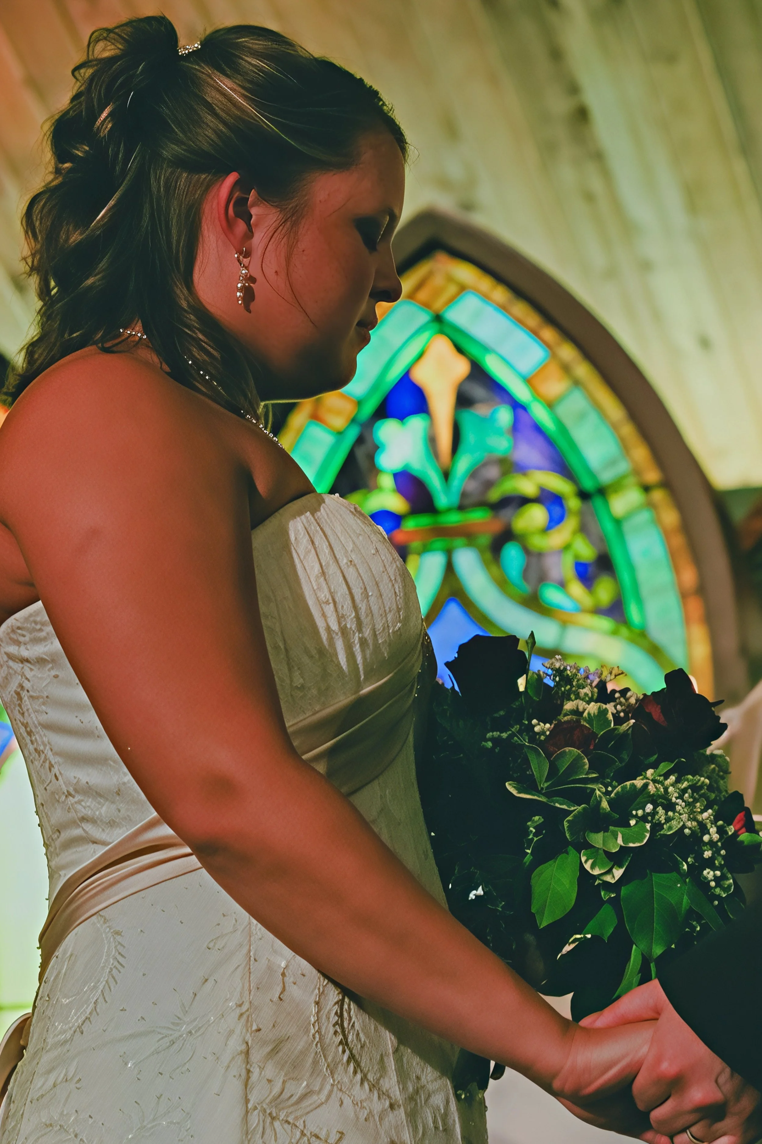A woman dressed in a white wedding gown holding a bouquet of flowers inside a church with stained glass windows.