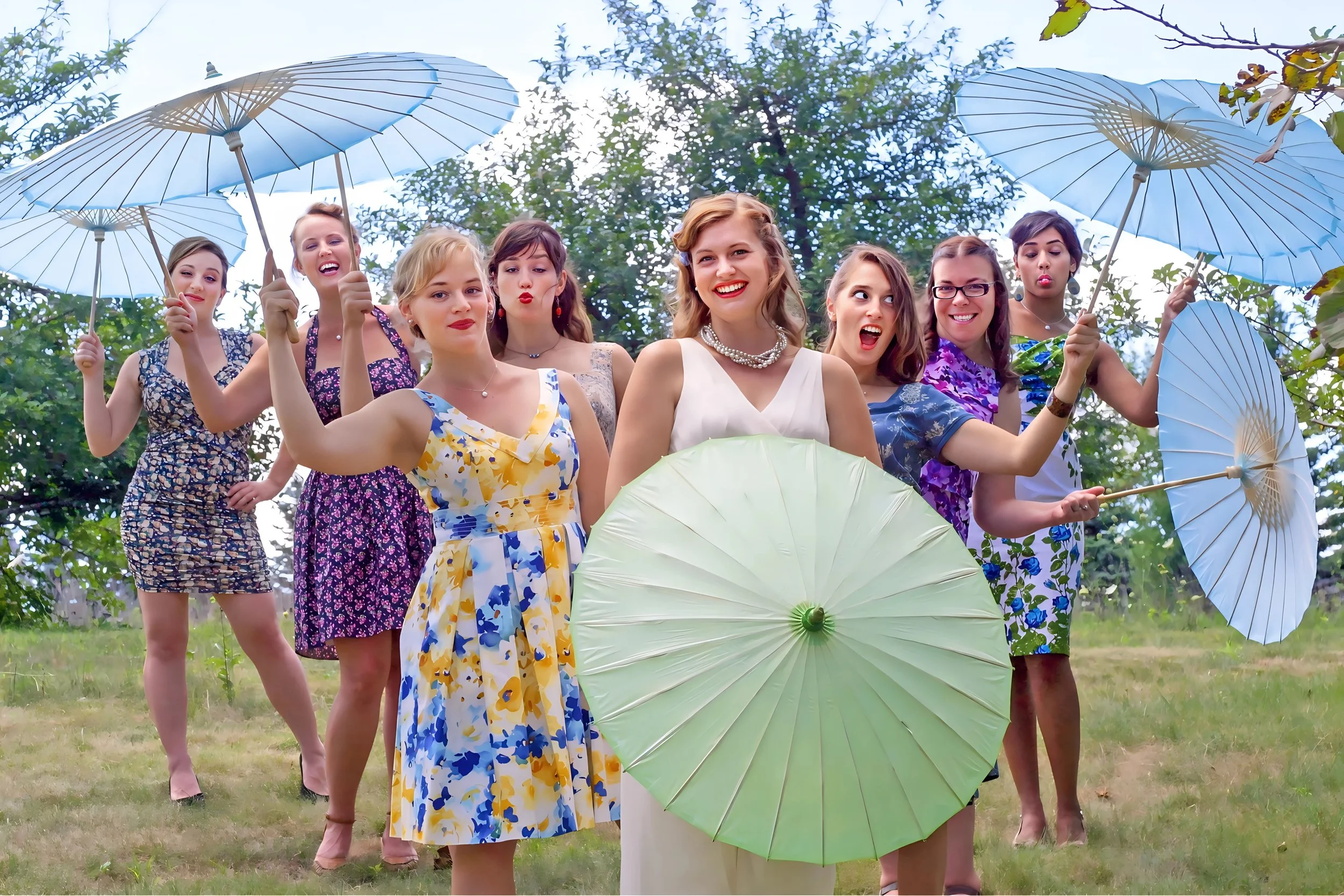 Group of women in floral dresses holding parasols outdoors, smiling and posing for a photo.