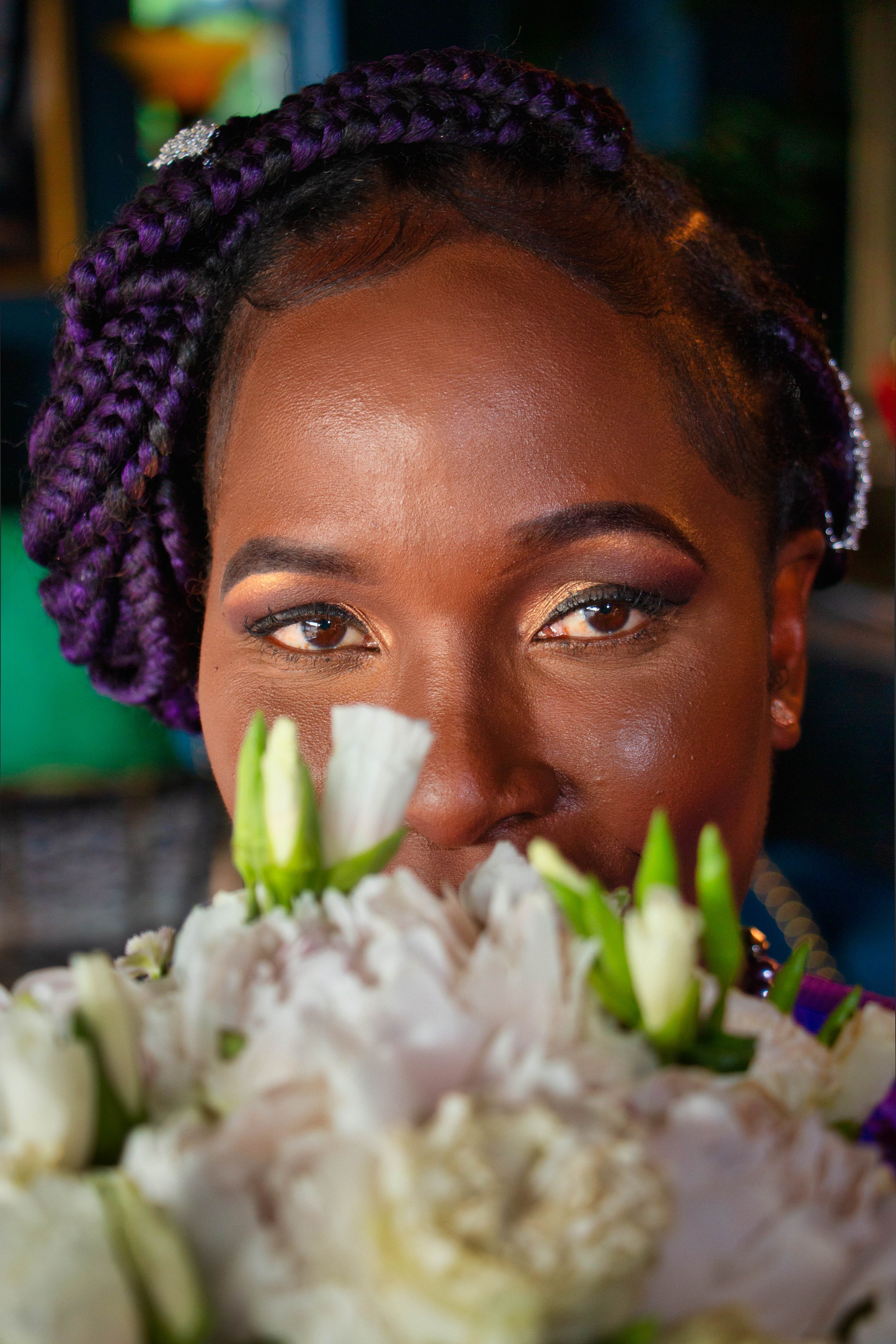 Close-up of a woman with braided purple hair and makeup, standing behind a bouquet of white flowers.