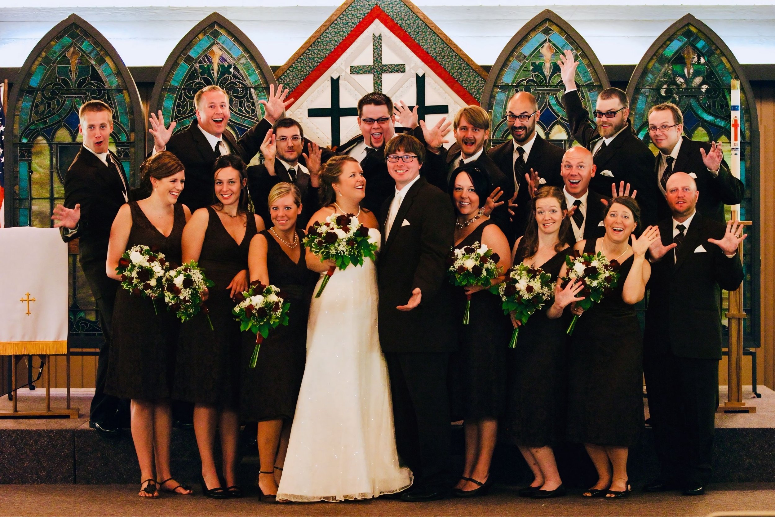 A wedding celebration with a group of people in a church, with the bride and groom in the center, surrounded by bridesmaids and groomsmen, all smiling and raising their hands.