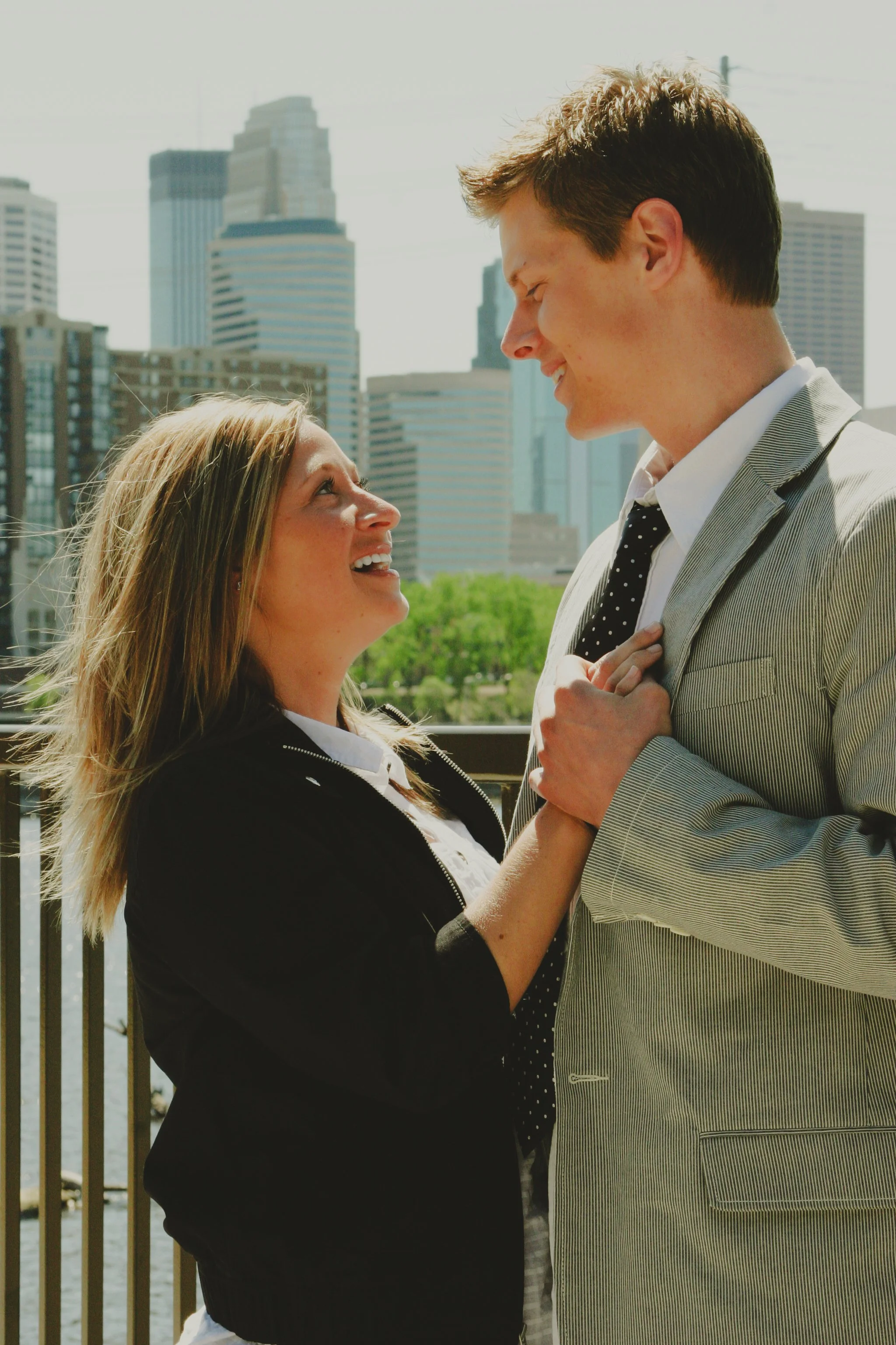 A man and woman smiling and holding hands in an outdoor urban setting with tall buildings in the background.
