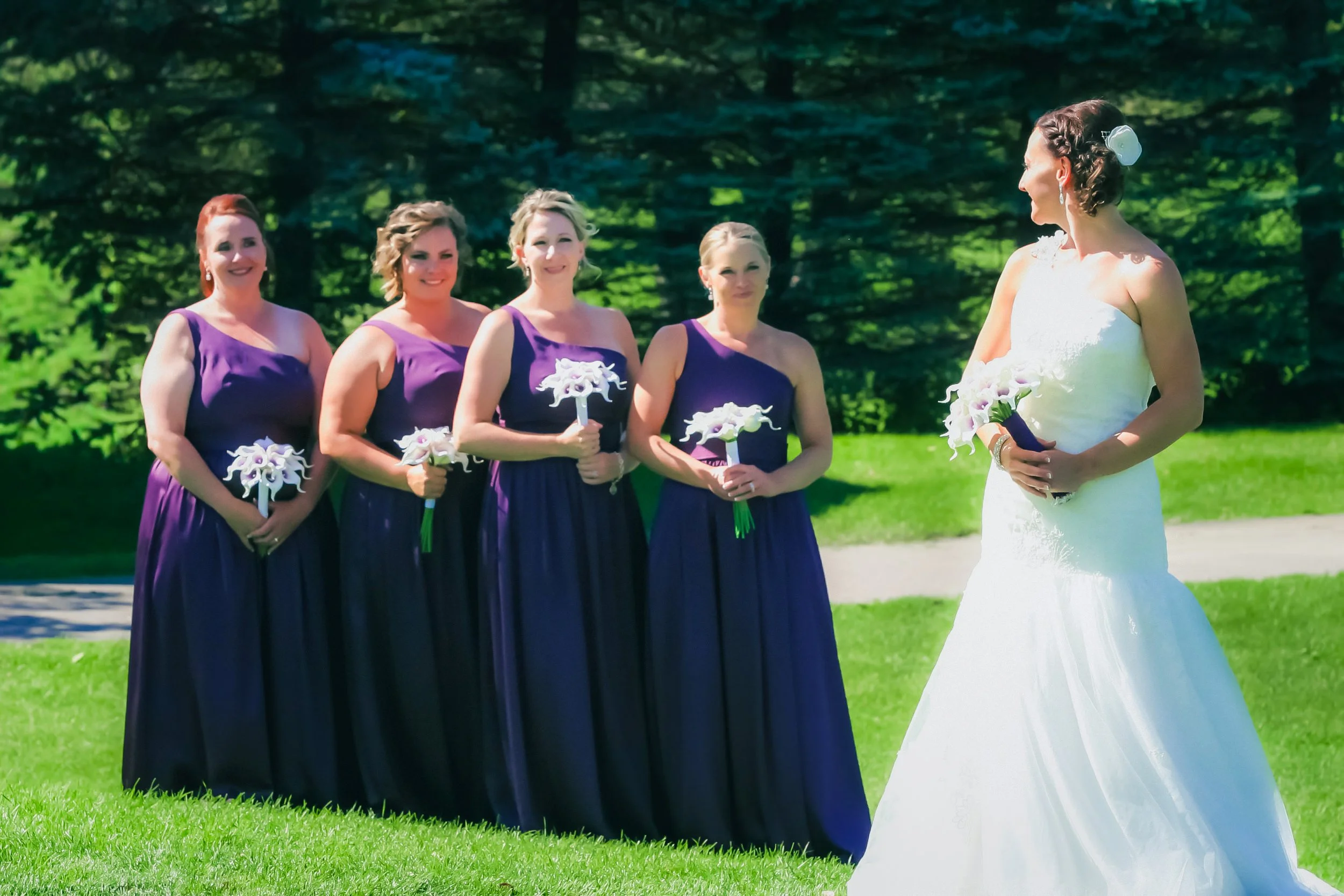 A bride in a white wedding dress holding a bouquet stands facing four bridesmaids in purple dresses, each holding a bouquet, outdoors on a sunny day with green trees and grass in the background.