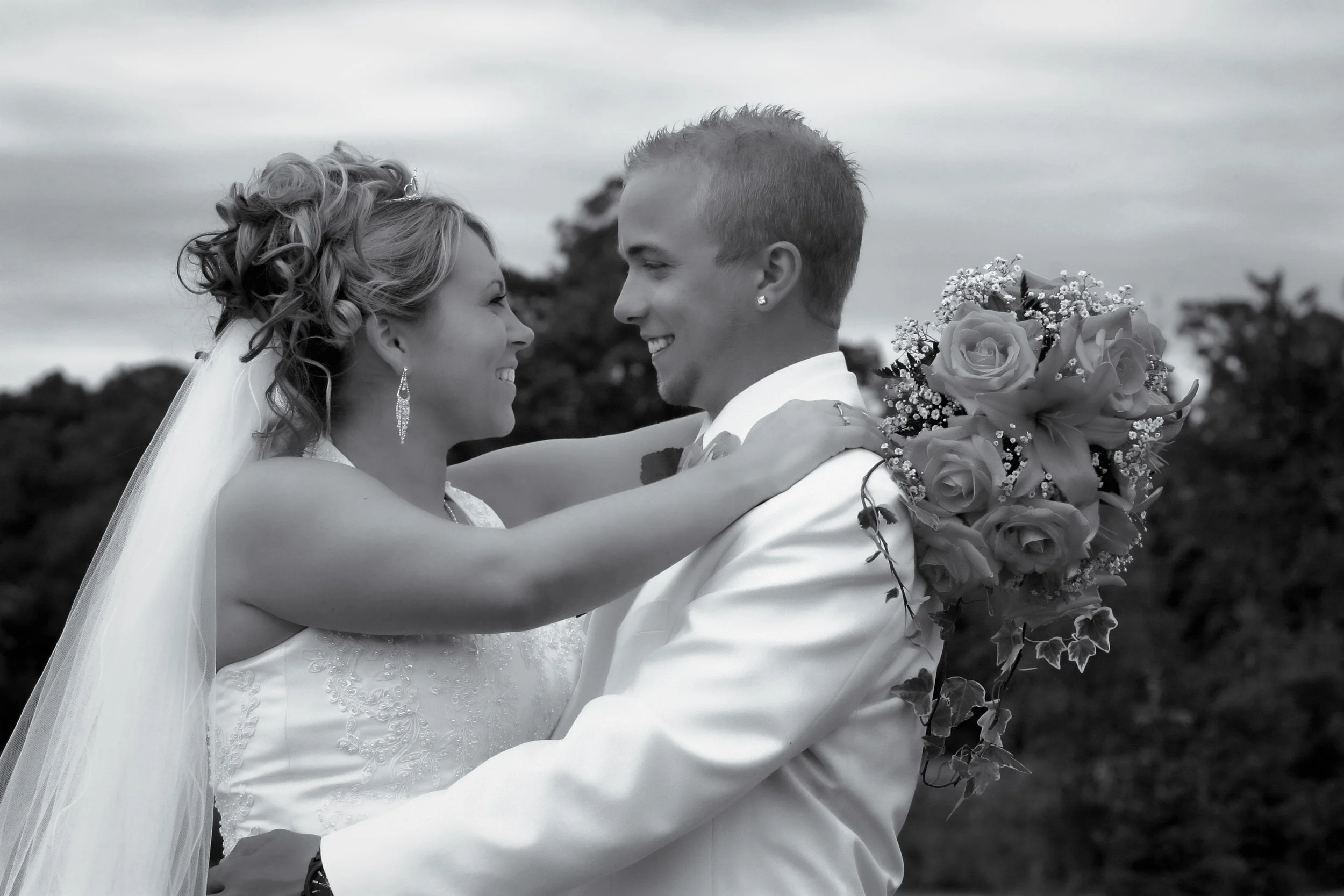 A bride and groom smiling at each other, embracing outdoors, with the bride holding a large bouquet of roses and wearing a wedding dress, veil, and earrings, in a black-and-white photo.
