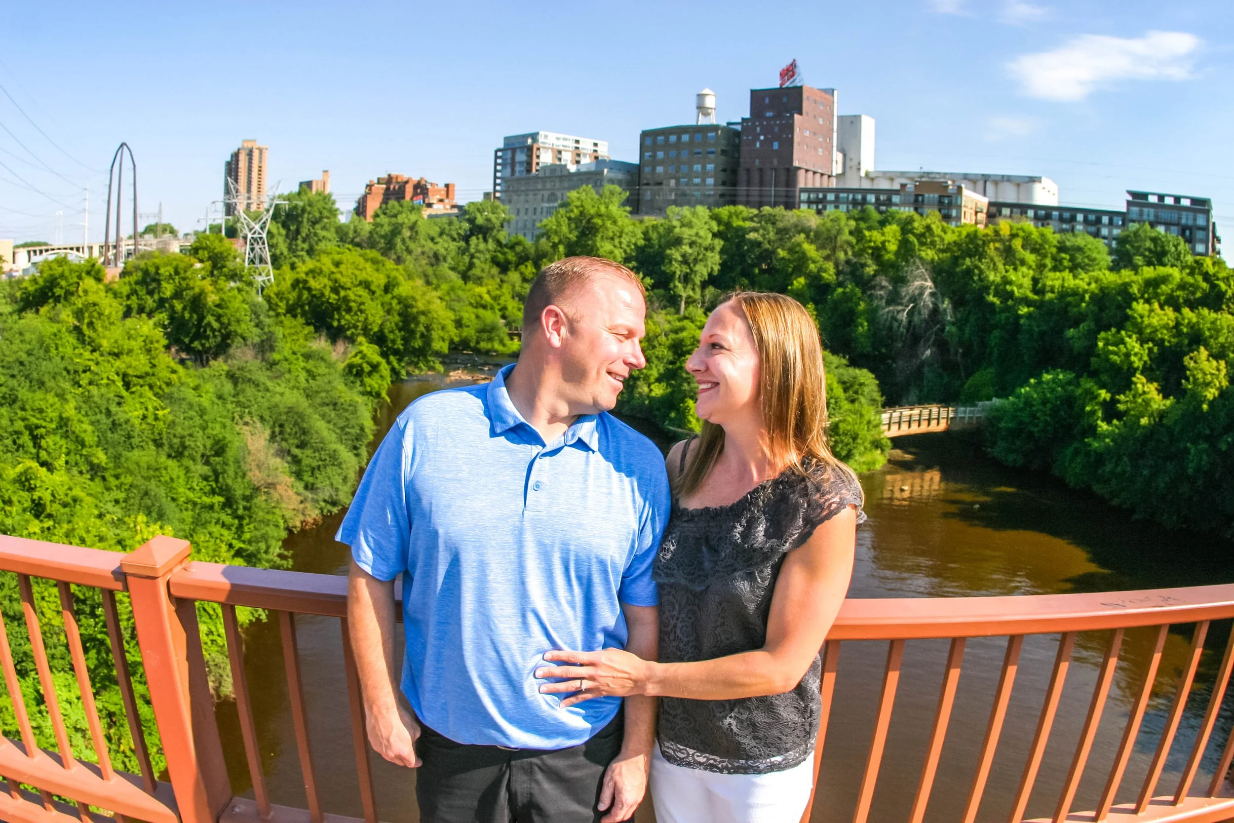 A smiling couple standing on a bridge with a river, green trees, and an urban skyline in the background on a sunny day.