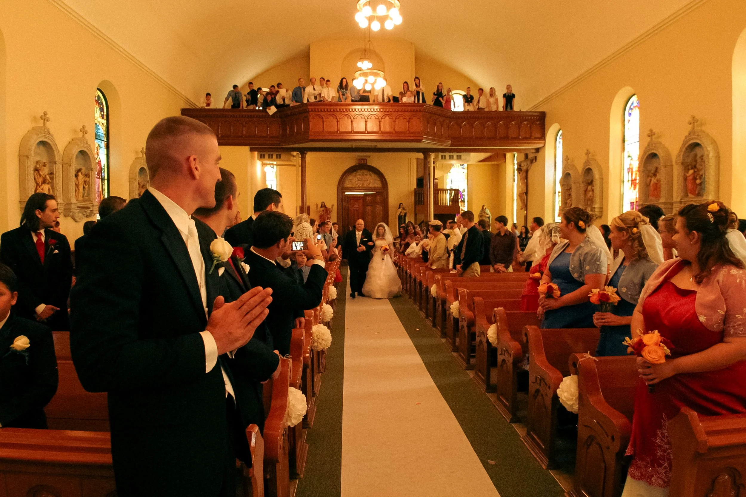 A wedding ceremony in a church with a bride and groom walking down the aisle, surrounded by guests dressed in formal attire, some taking photos, warm lighting, stained glass windows, and religious artwork on the walls.