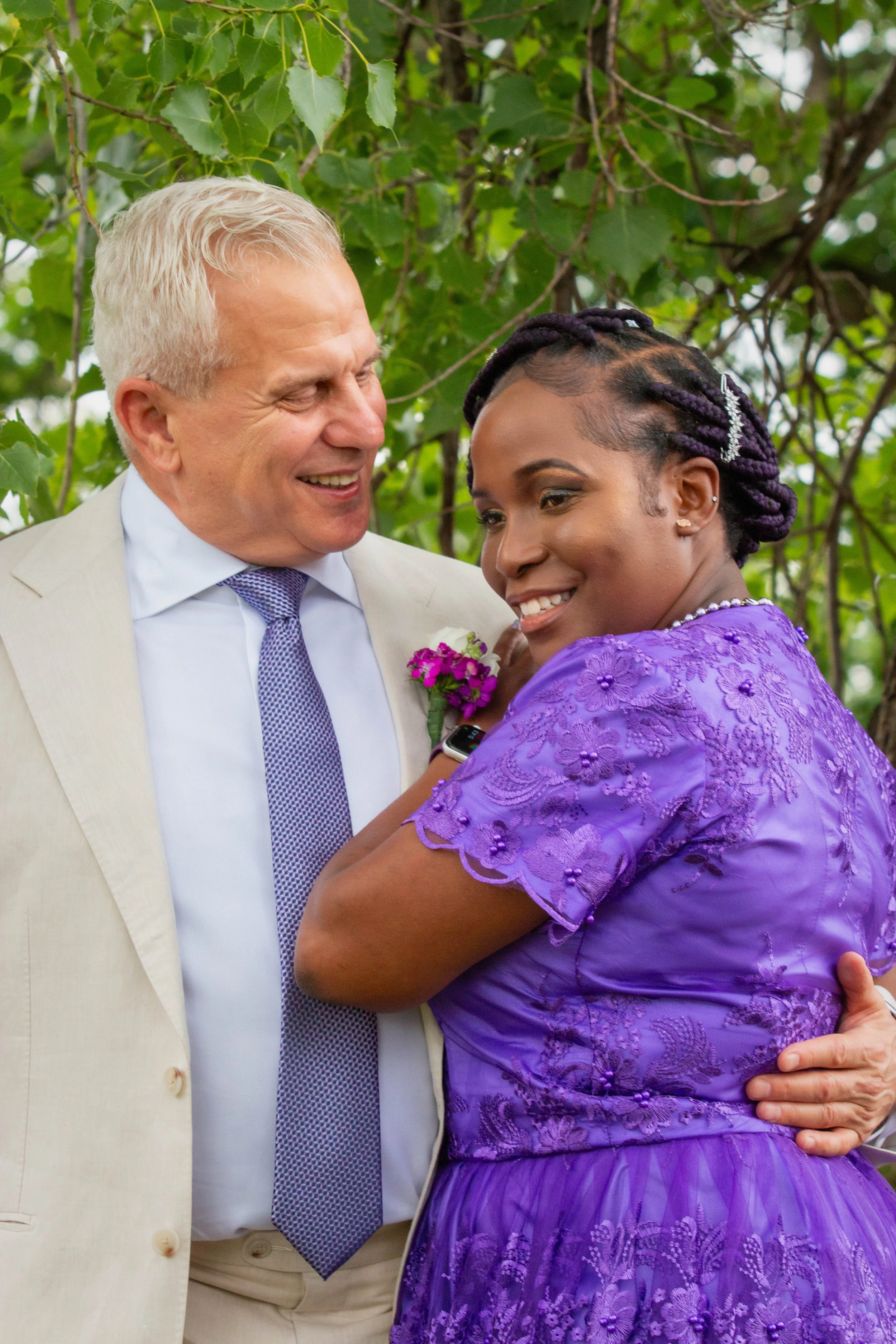 A man and woman sharing a joyful moment outdoors, the man wearing a light-colored suit and tie with a pink boutonniere, and the woman wearing a purple dress with embroidered floral patterns, both smiling and embracing.