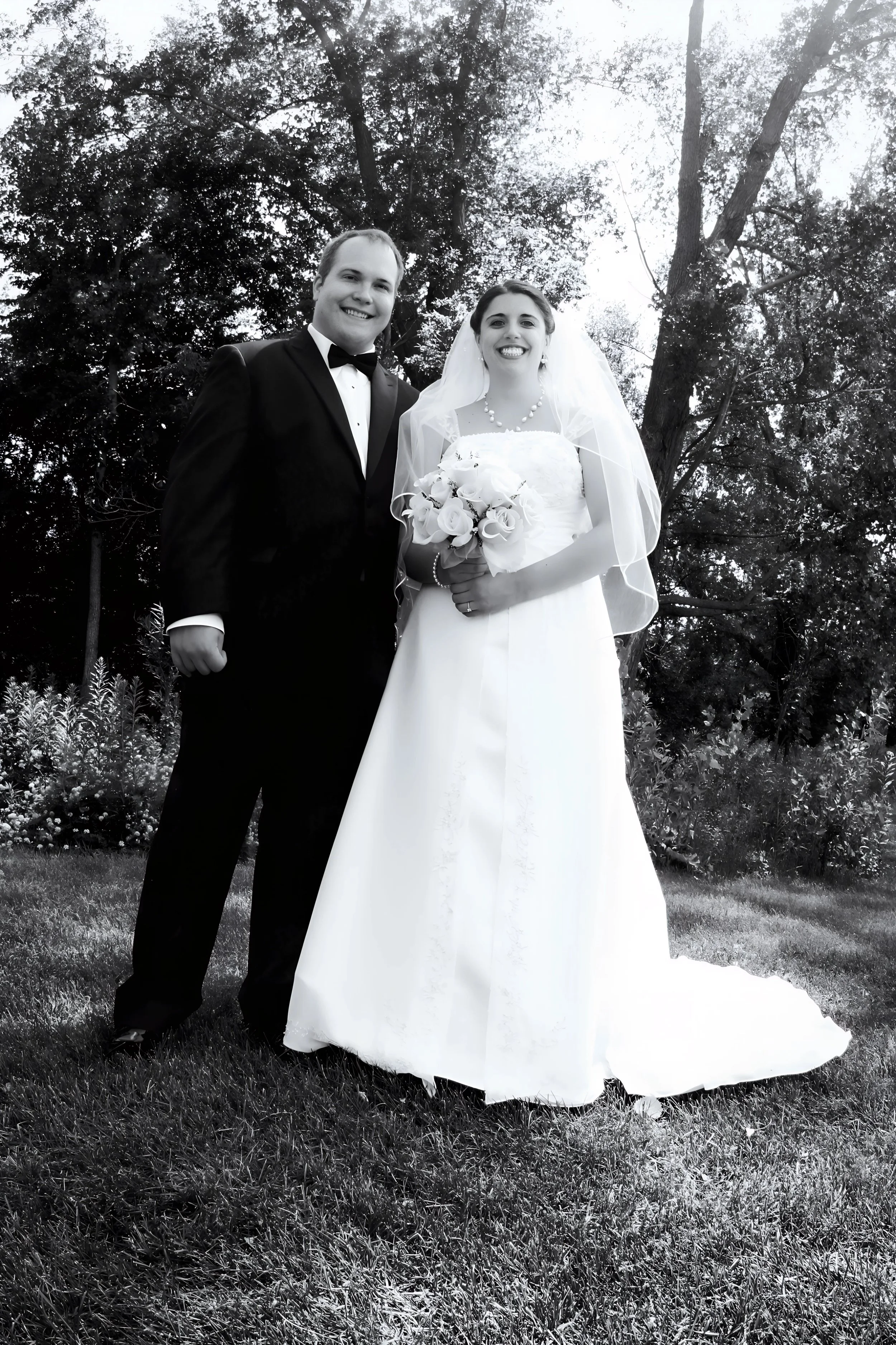 Black and white photo of a bride and groom standing outdoors on grass, smiling. The groom wears a tuxedo, and the bride wears a wedding dress and veil, holding a bouquet of flowers. Trees are in the background.