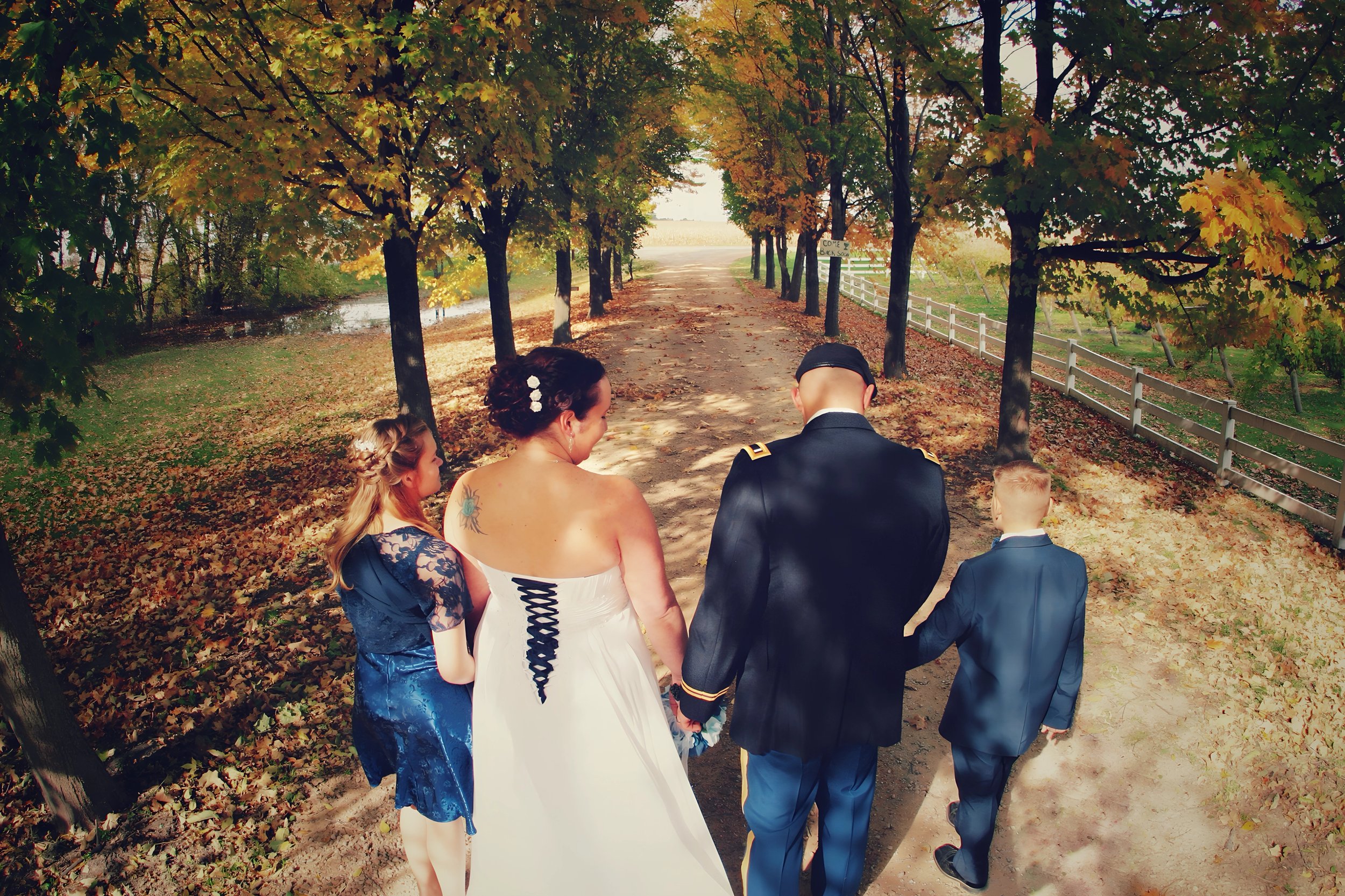 A wedding couple holding hands walking on a dirt path with two children in formal attire, under a canopy of autumn trees.