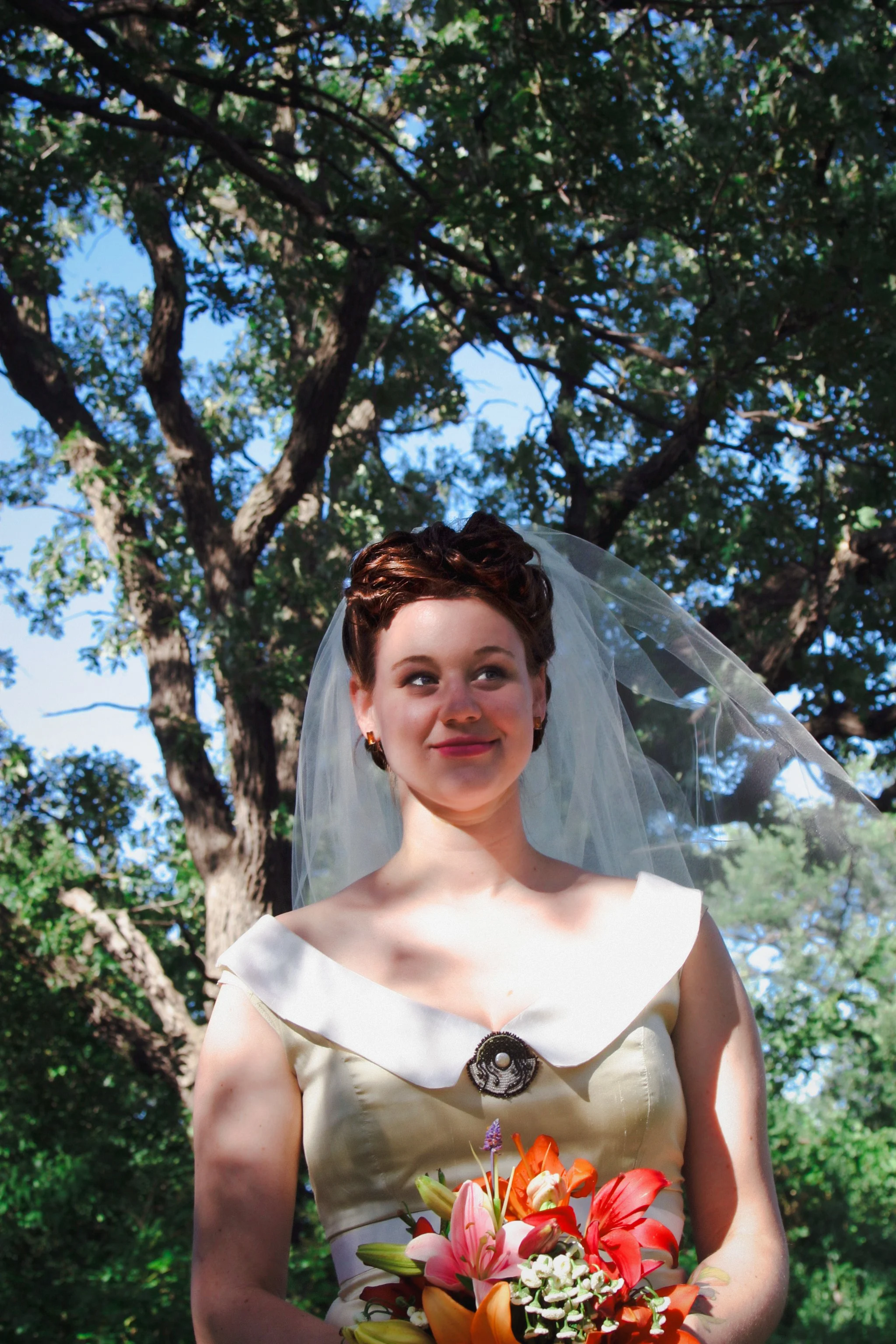 A woman in a vintage wedding dress with a veil holding a bouquet of colorful lilies and other flowers standing outdoors under a large tree.