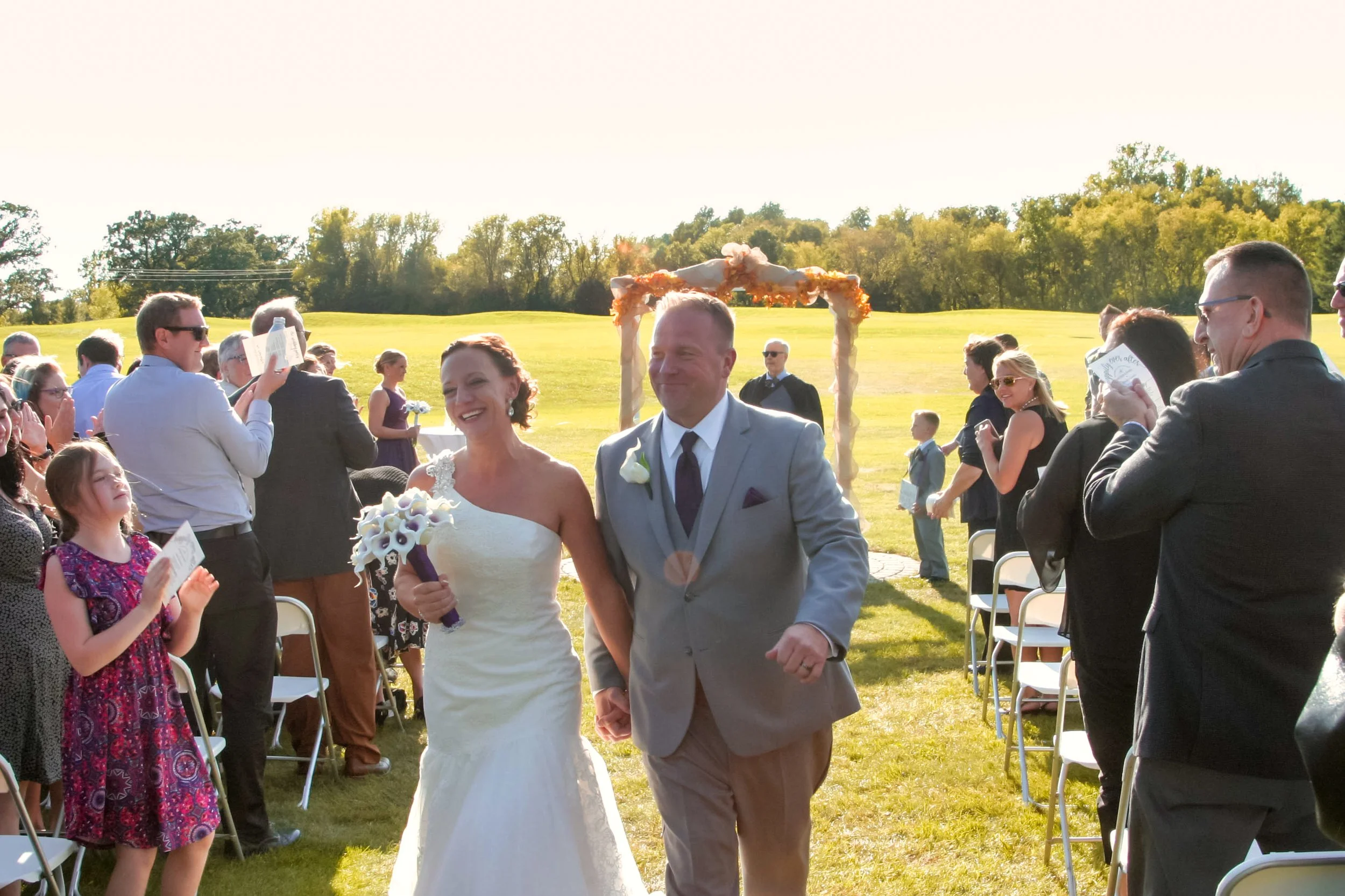 Bride and groom walking down aisle outdoors at their wedding ceremony, surrounded by guests, with a decorated arch and green landscape in the background.