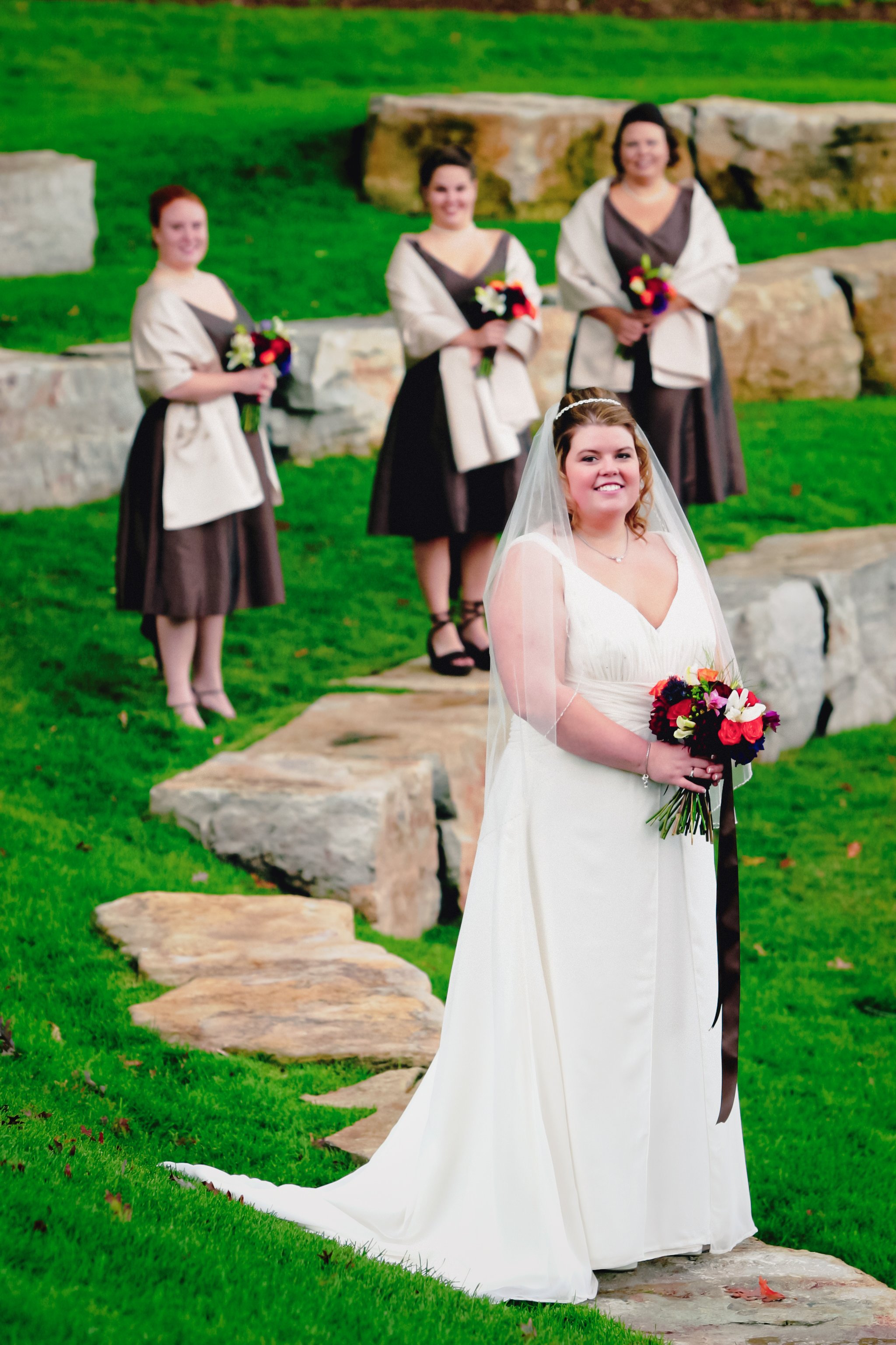 A bride in a white wedding dress holding a bouquet of red, white, and purple flowers in front of her, standing on a stone pathway with green grass. Behind her, three women are standing on a staircase made of stones, holding smaller bouquets, dressed 