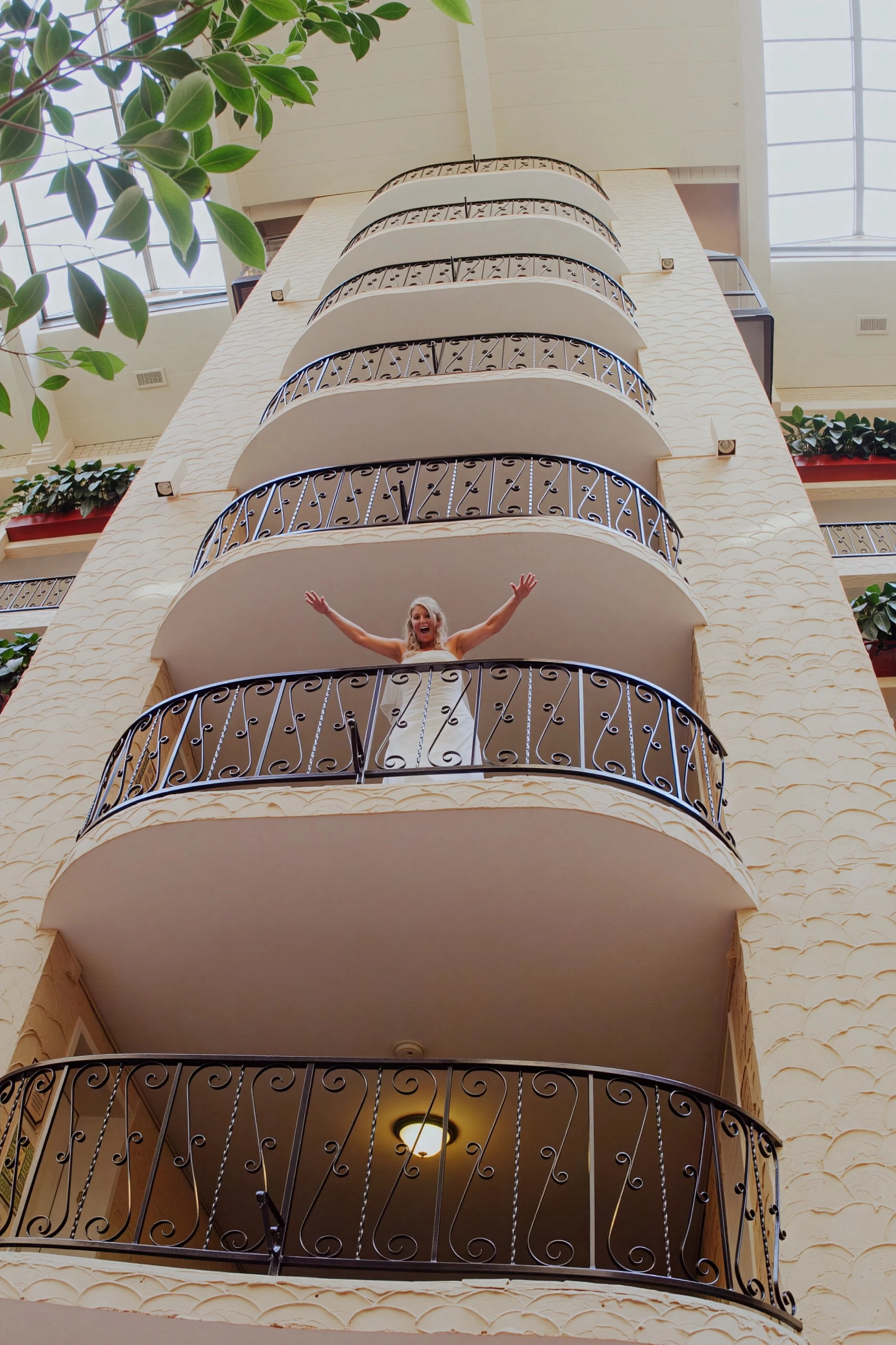 A woman in a white dress standing on a hotel balcony with her arms raised, smiling, with multiple balconies and green plants surrounding her.