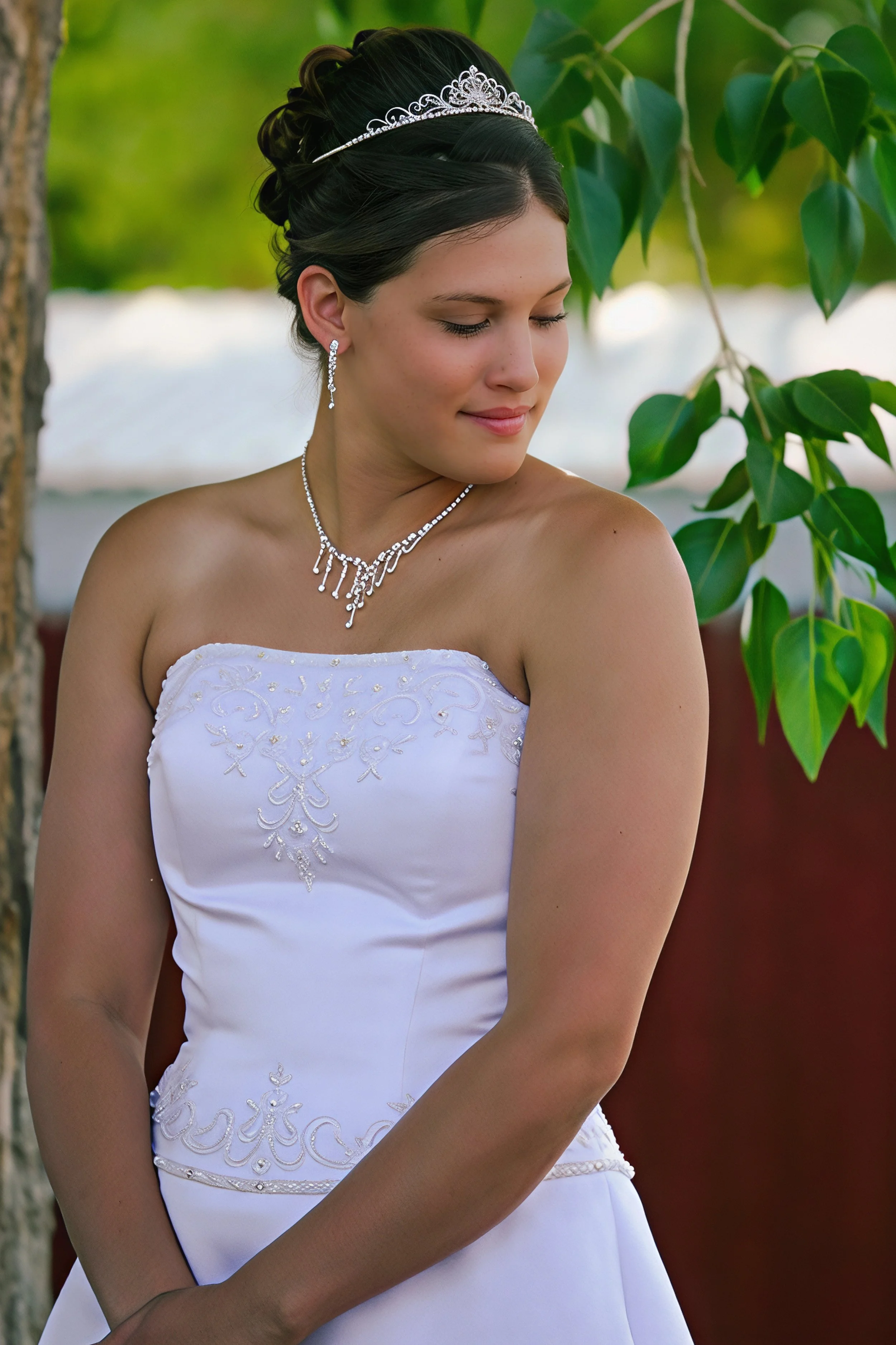 A woman dressed in a white wedding gown with intricate embroidery, wearing a silver tiara and jewelry, standing outdoors near green foliage.