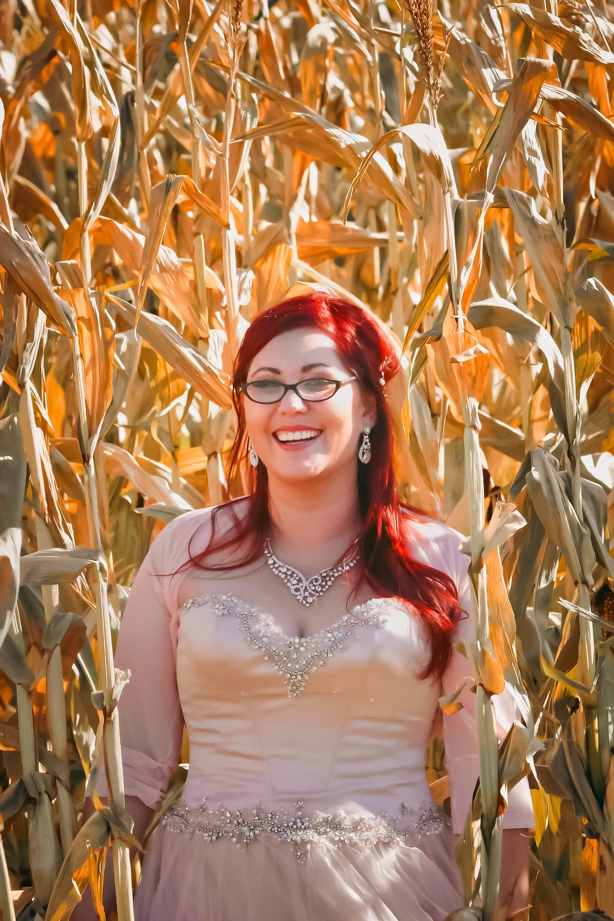 A woman with red hair and glasses in a wedding dress smiling in a cornfield