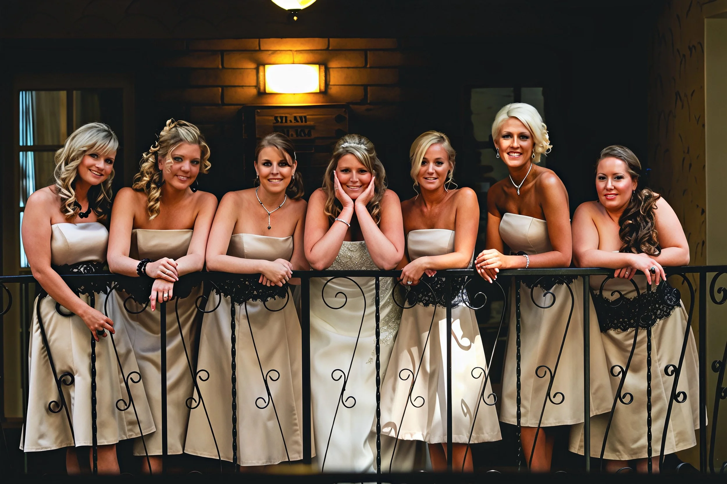 Seven women dressed in elegant matching beige and black dresses, posing together behind a decorative black metal railing in an indoor setting, likely at a wedding or formal event.