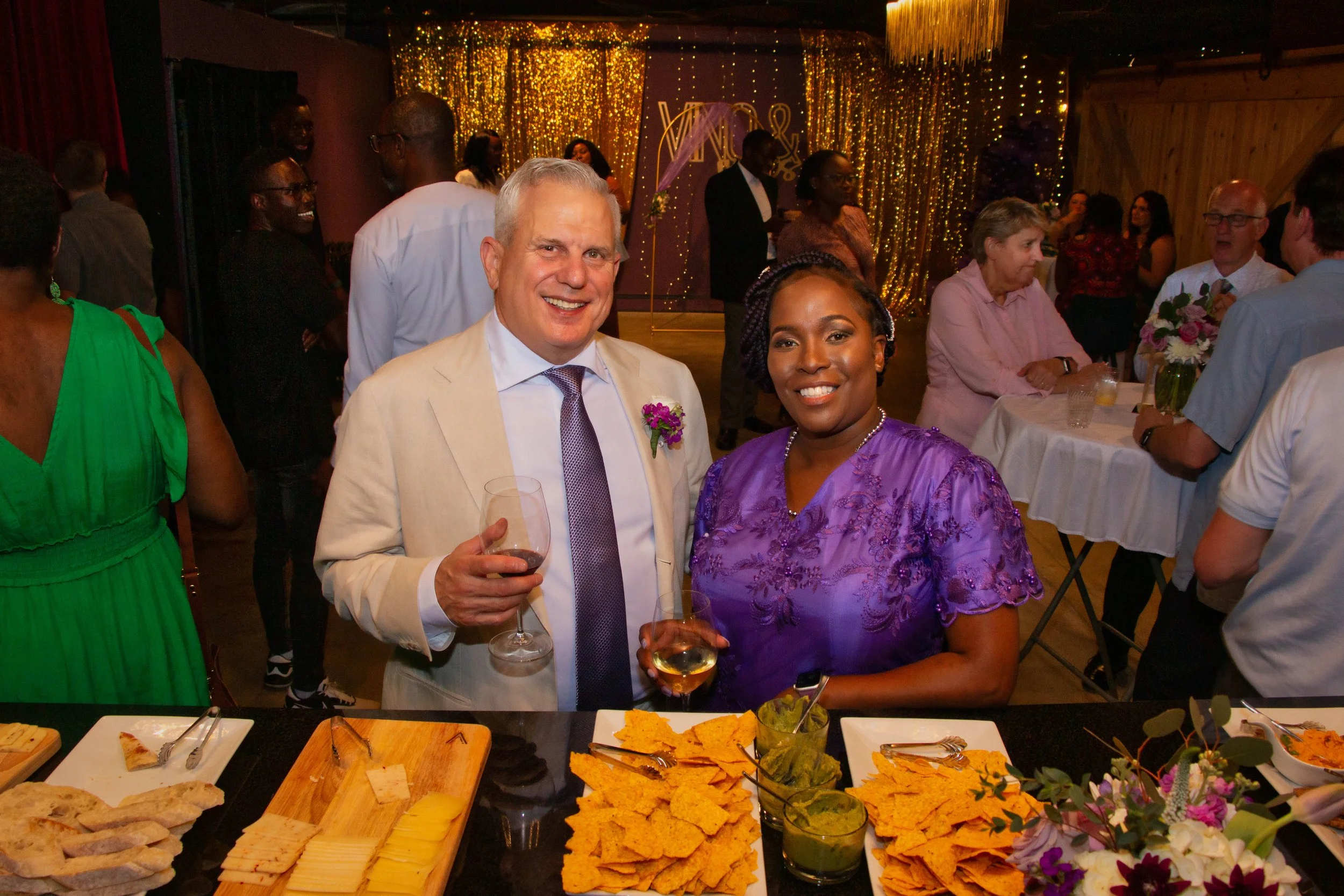 Two people at a celebration or wedding reception, smiling and holding drinks, standing in front of a table with snacks and food, with other guests in the background and a gold sequin backdrop.