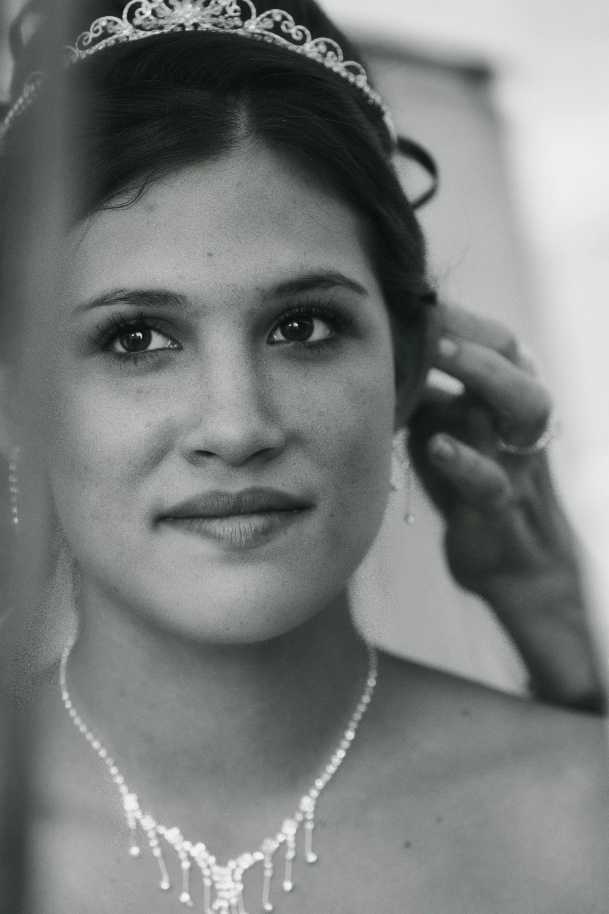 Black and white close-up photo of a bride adjusting her earring, wearing a tiara and pearl necklace, with a soft smile and expressive eyes.