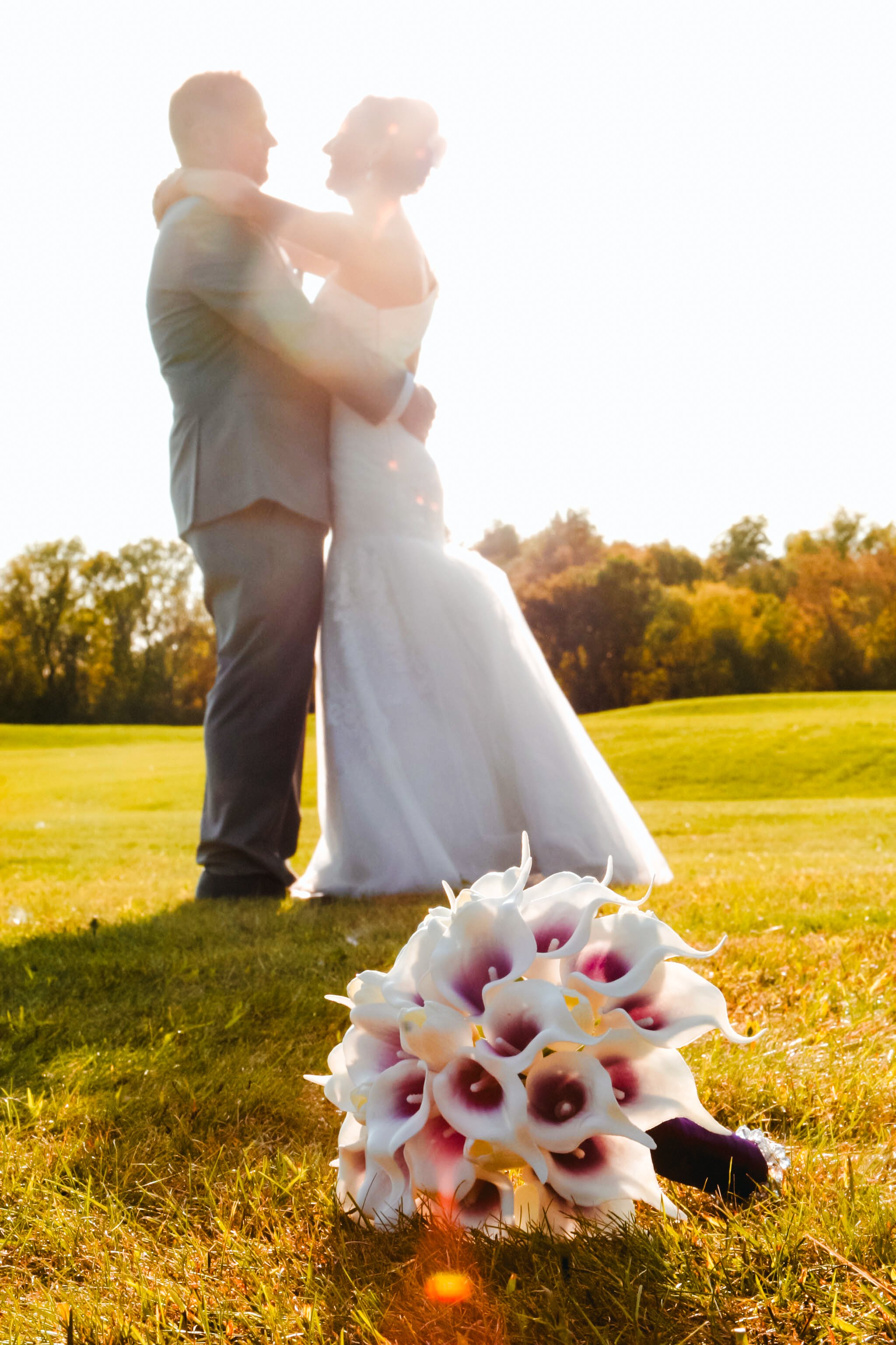 A bride and groom are embracing and gazing at each other outdoors during sunset, with a bouquet of calla lilies in the foreground and yellow and orange trees in the background.