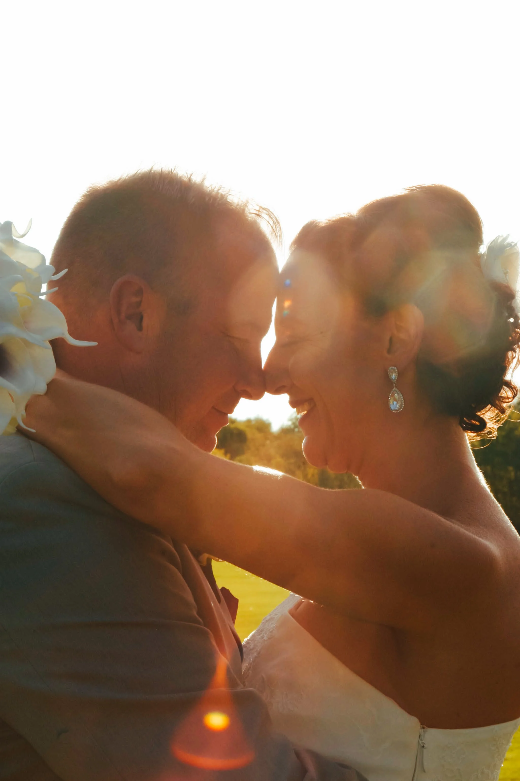 A couple on their wedding day, embracing and about to kiss, with the sun setting behind them, creating a warm glow and lens flare.