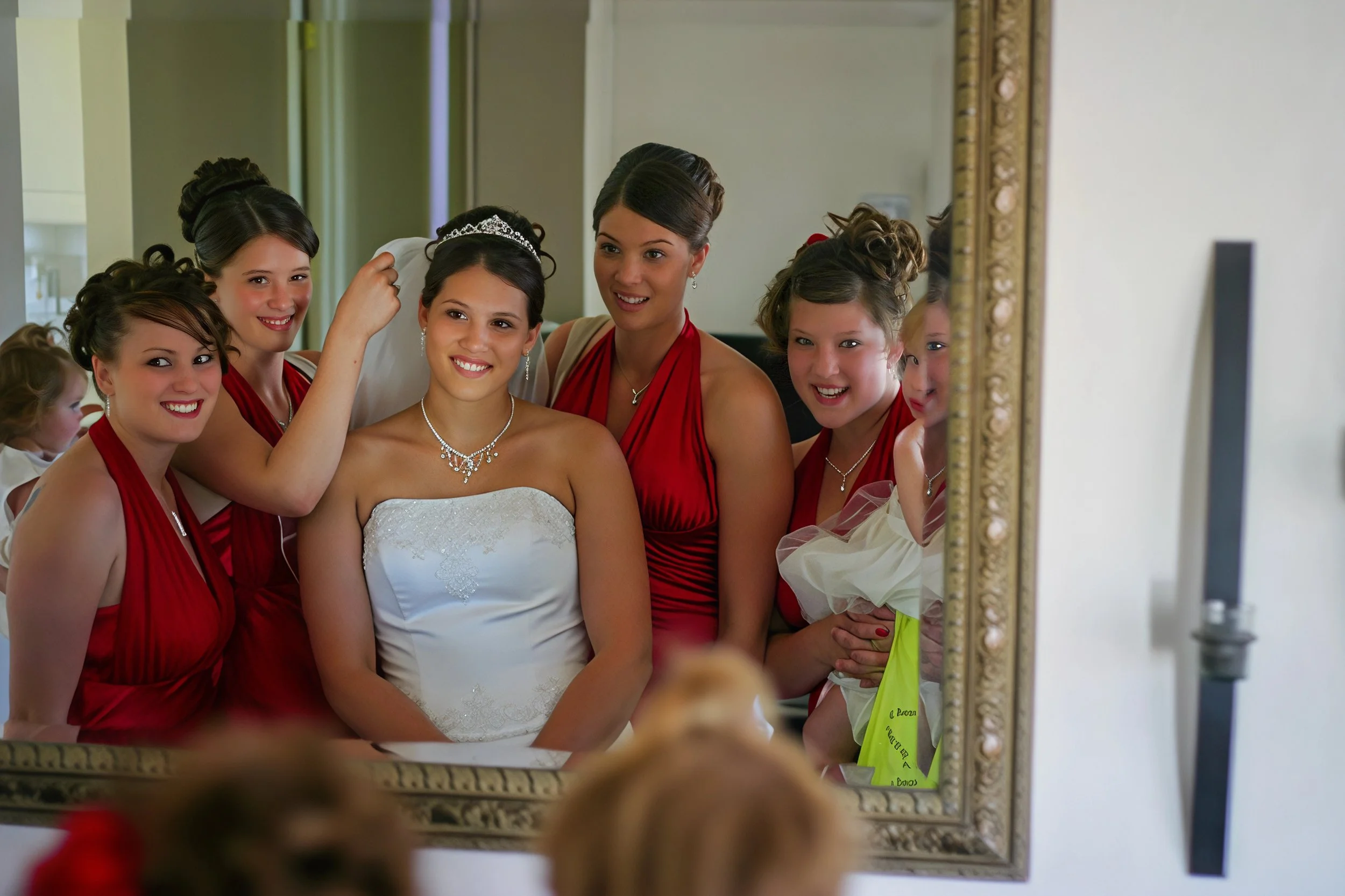 Bride in white dress and tiara taking a selfie with her bridesmaids in red dresses and young flower girl in white, all smiling and posing in front of a mirror.