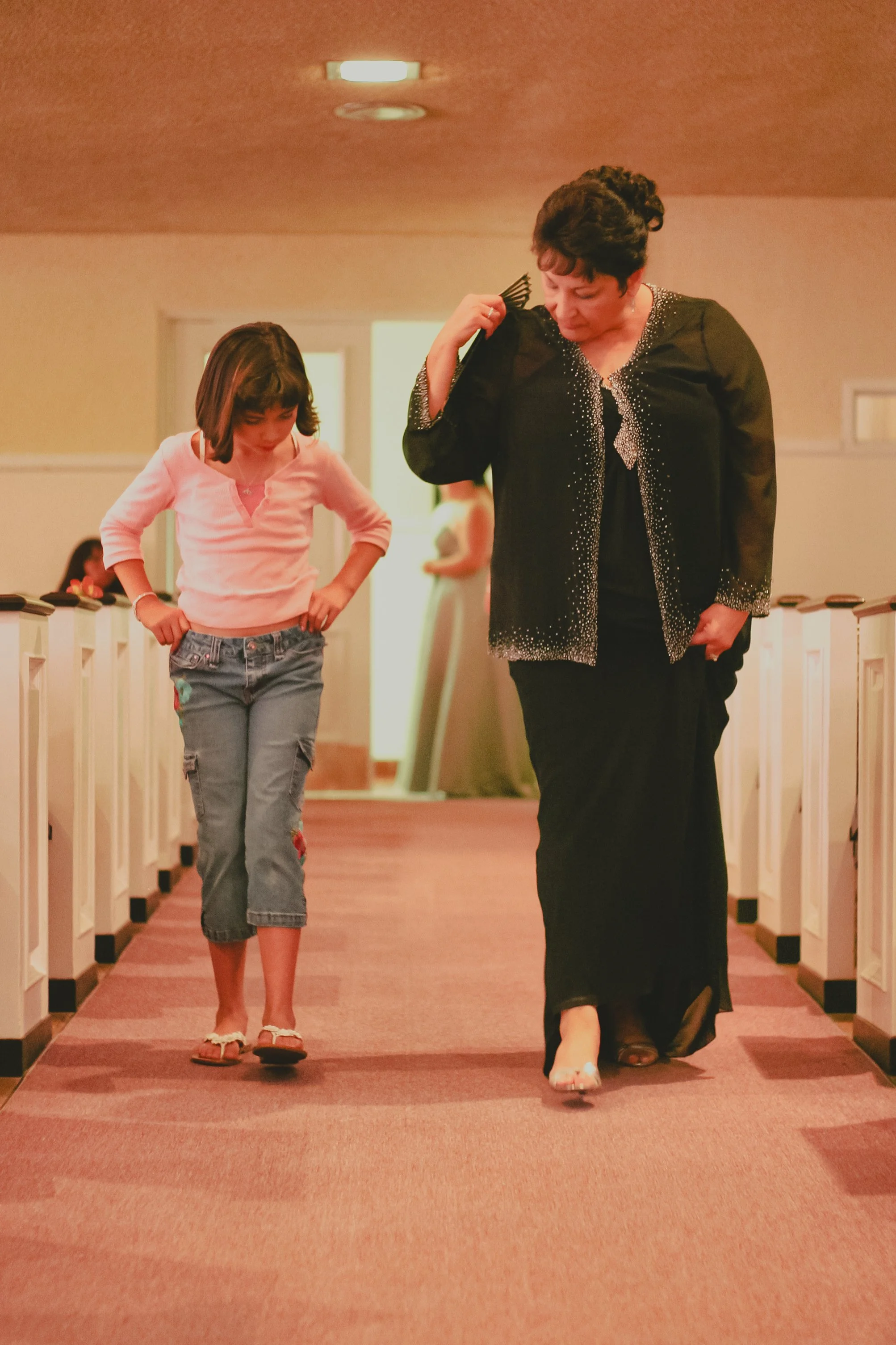 A woman and a young girl walking down a church aisle, both looking down, with the woman holding a fan over her shoulder.