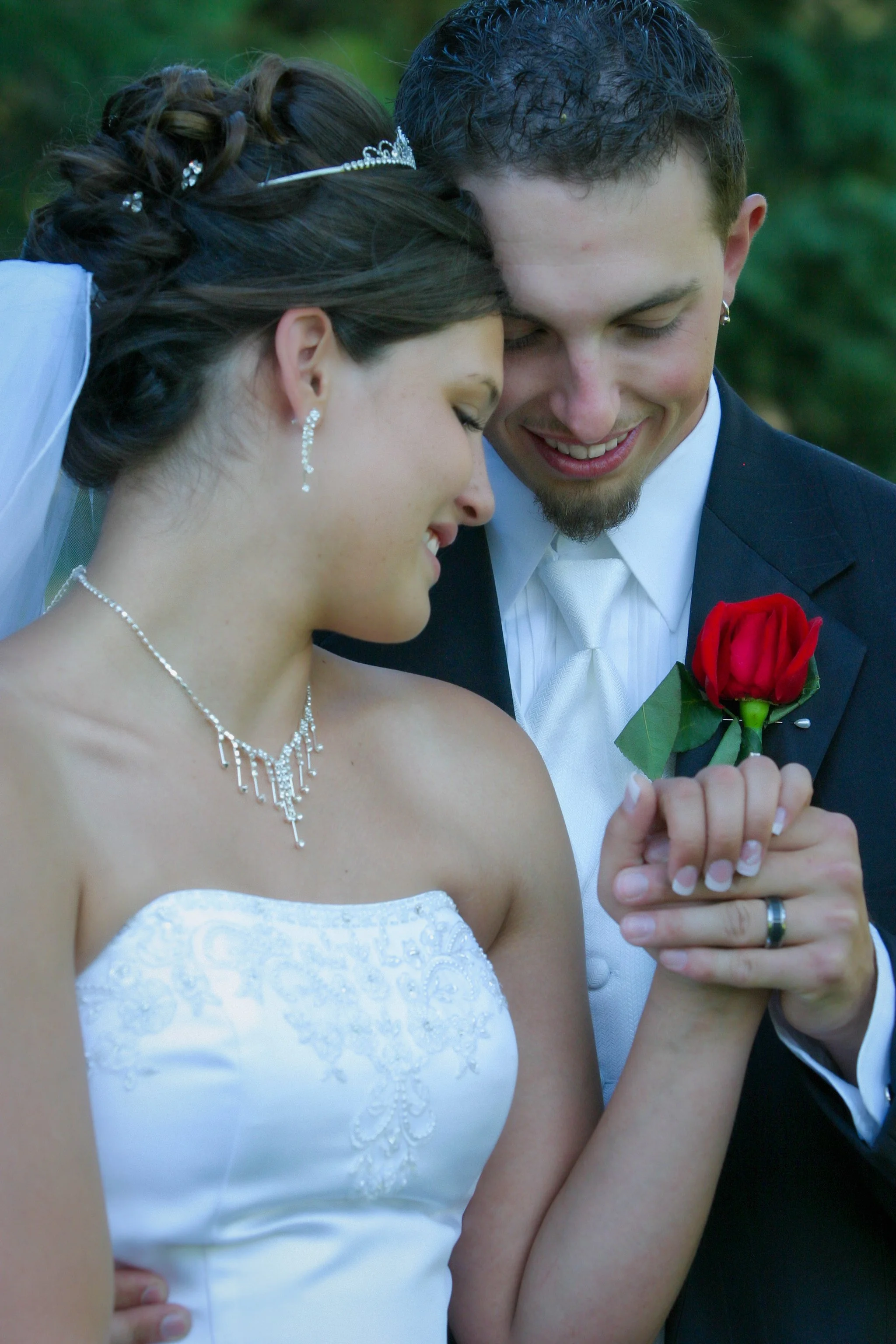 A bride and groom sharing a tender moment, holding a red rose together, with the bride wearing a white wedding dress and jewelry, and the groom in a black tuxedo, outdoors with greenery in the background.
