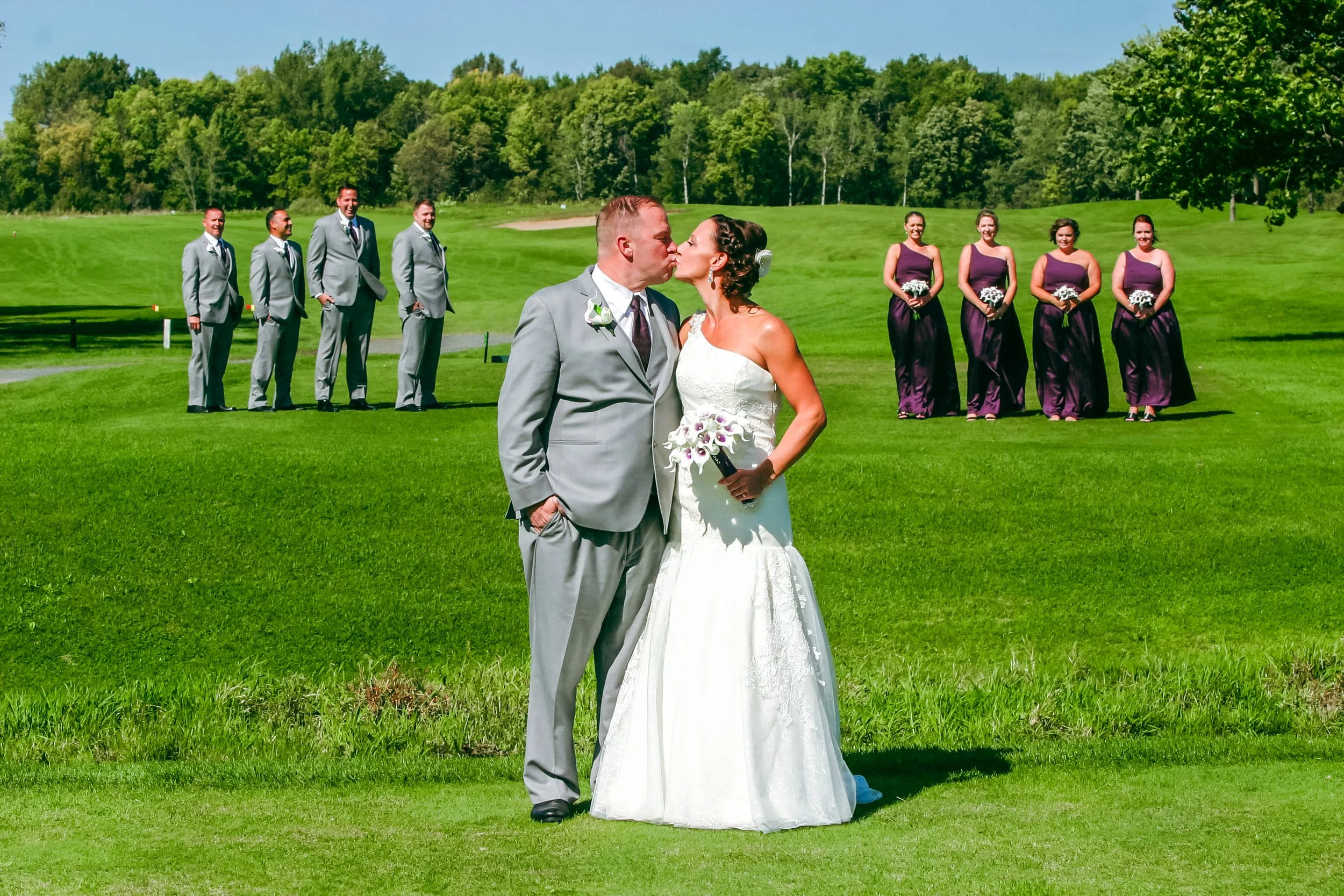 A newlywed couple sharing a kiss on a golf course with the wedding party in the background.