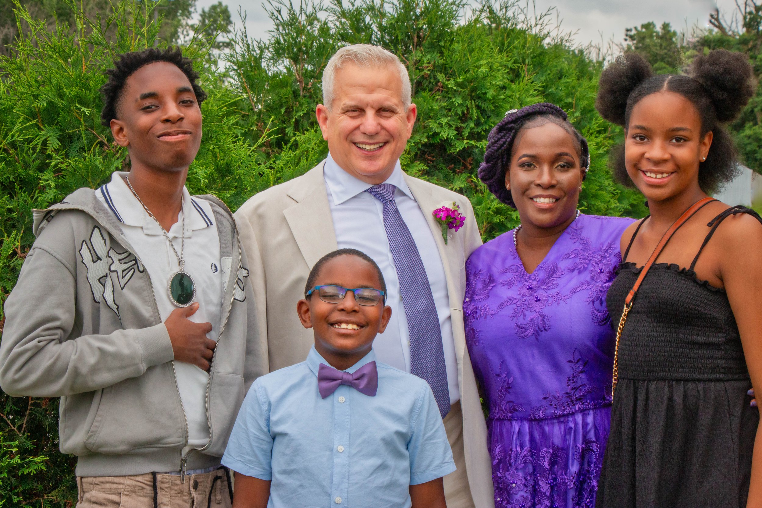 Group of five people, including a man and four children, smiling outdoors with greenery in the background.
